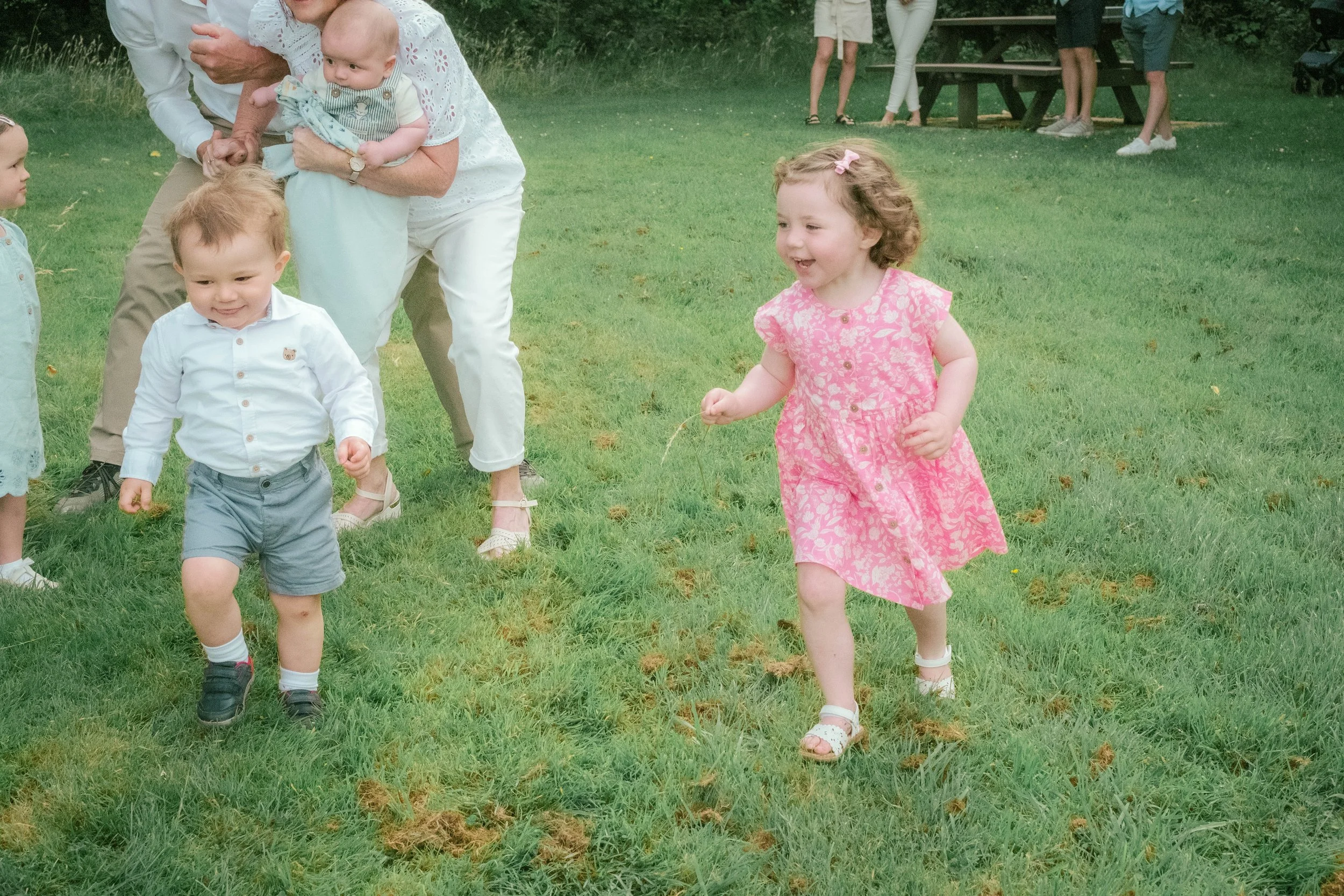 A group of children and adults outdoors on a grassy field, with a young girl in a pink dress running and smiling, and a boy in a white shirt and gray shorts jogging nearby.
