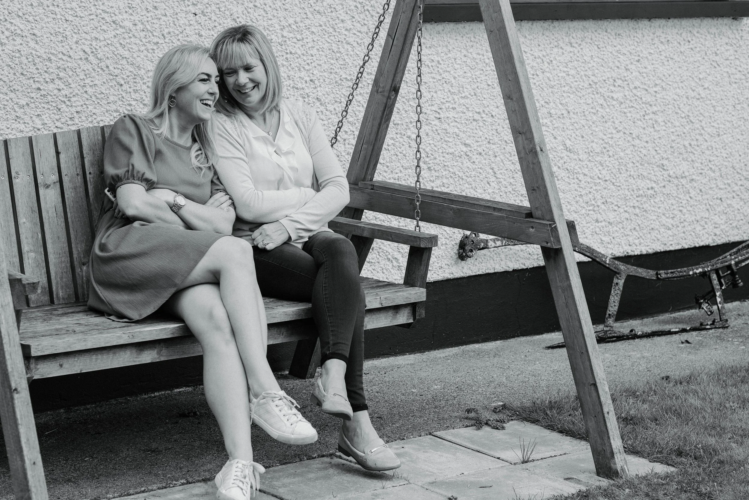 Two women sit on a wooden park bench next to a swing set, laughing and smiling together, outdoors in front of a textured wall.