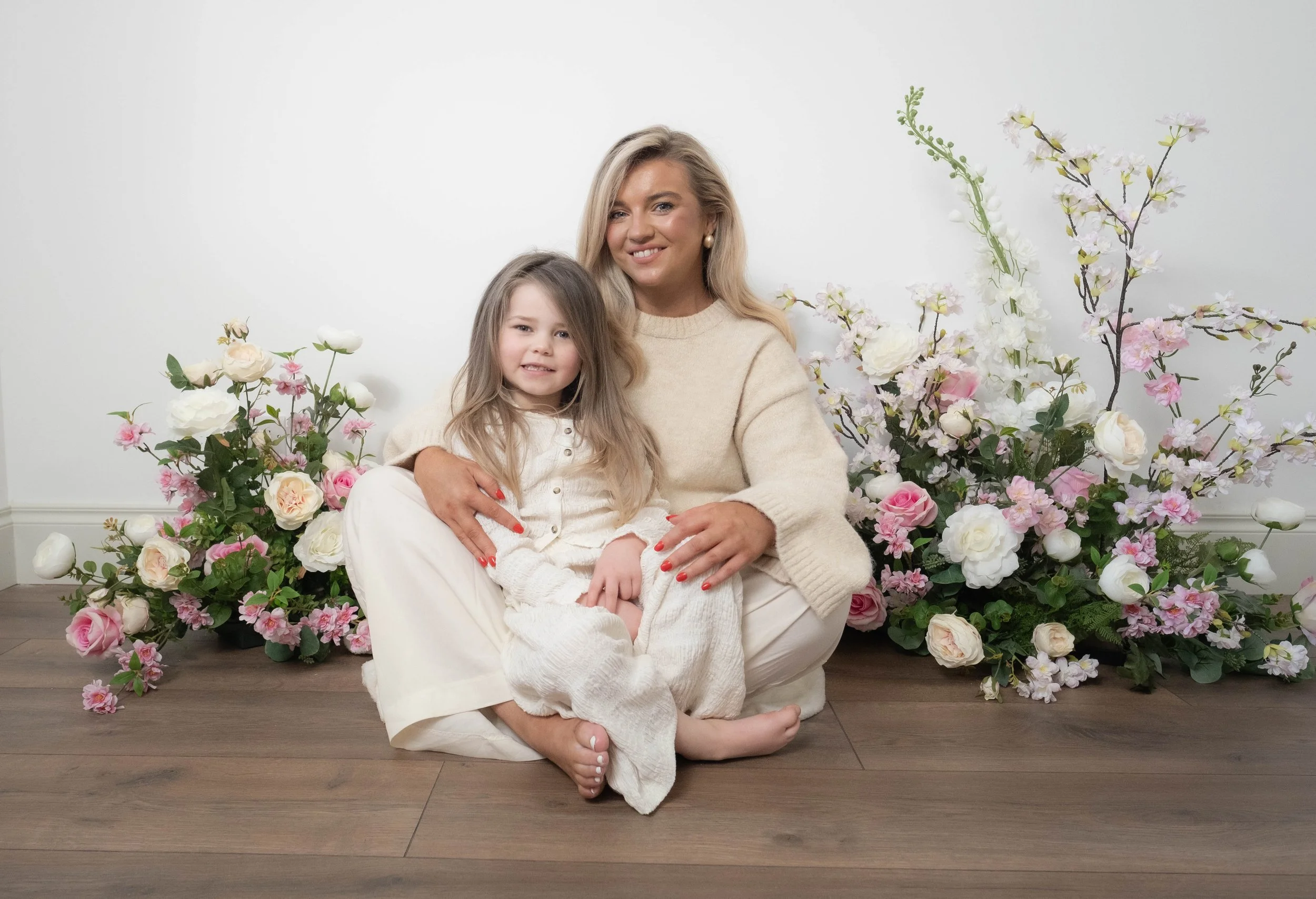 A woman and young girl sitting on the floor surrounded by pink and white flowers, with the woman holding the girl in her lap, both smiling at the camera.
