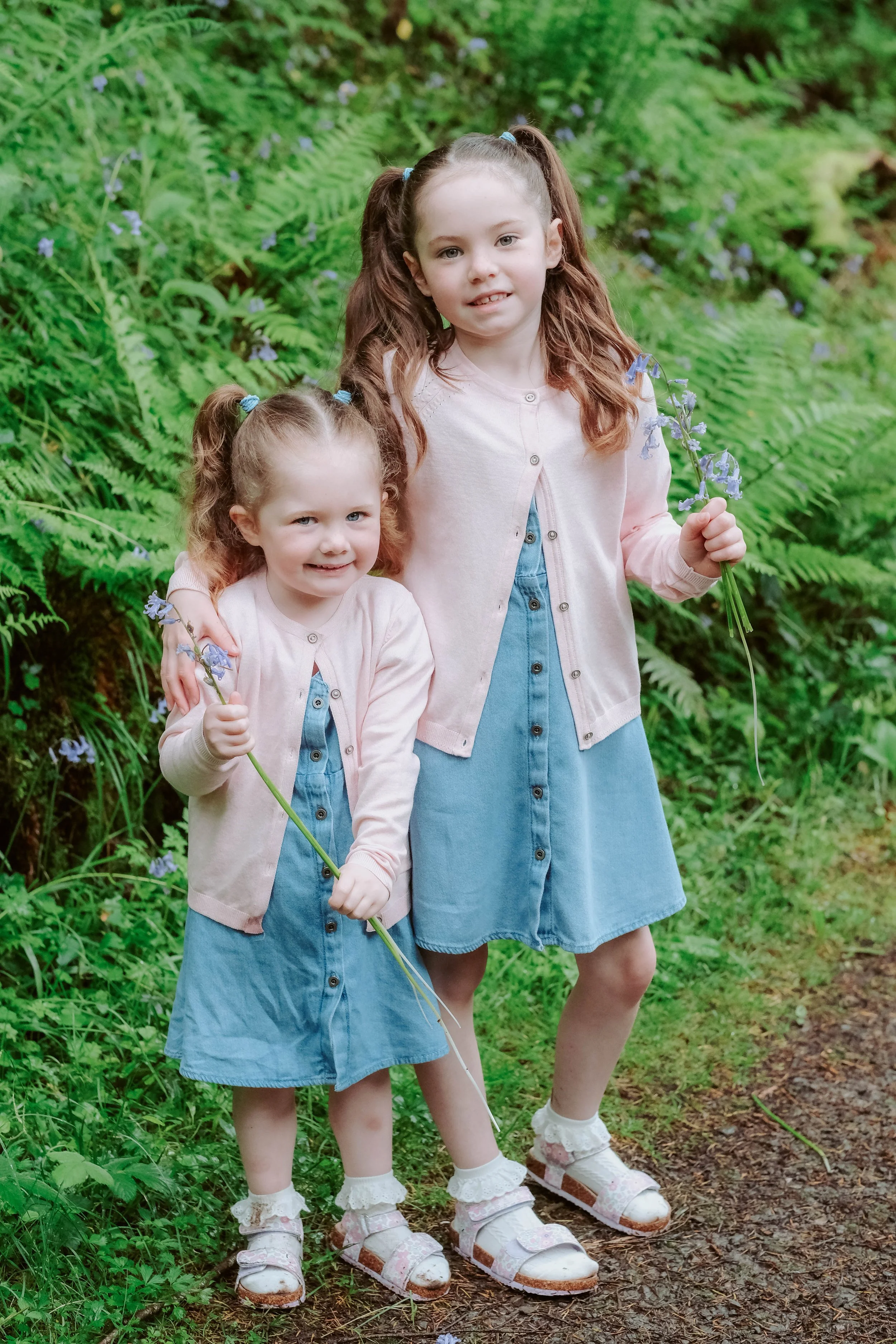 Two young girls with hair in pigtails standing on a forest trail, holding purple flowers, surrounded by green ferns.