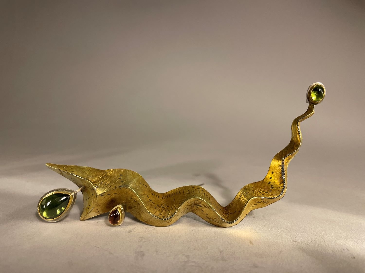 ‘River Origin’ Brooch (Swoop Series), 1993, materials 18k gold laminate, sterling, 24k Bezels, Tear drop Green Tourmaline, Tear
drop Garnet Oval Peridot, (13x5.5x2cm), $3000 Cdn.