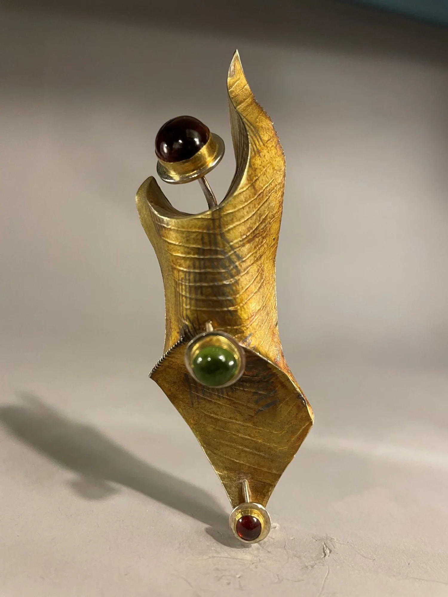 ‘Figure’ Brooch (Swoop Series), 1998, fusing, scoring and bending, soldering and construction, materials 18k gold laminate, sterling, 24k Bezels, Large Oval Garnet Top, Round Peridot, Round Garnet, (9.2x3x3 cm), $1800 Cdn.