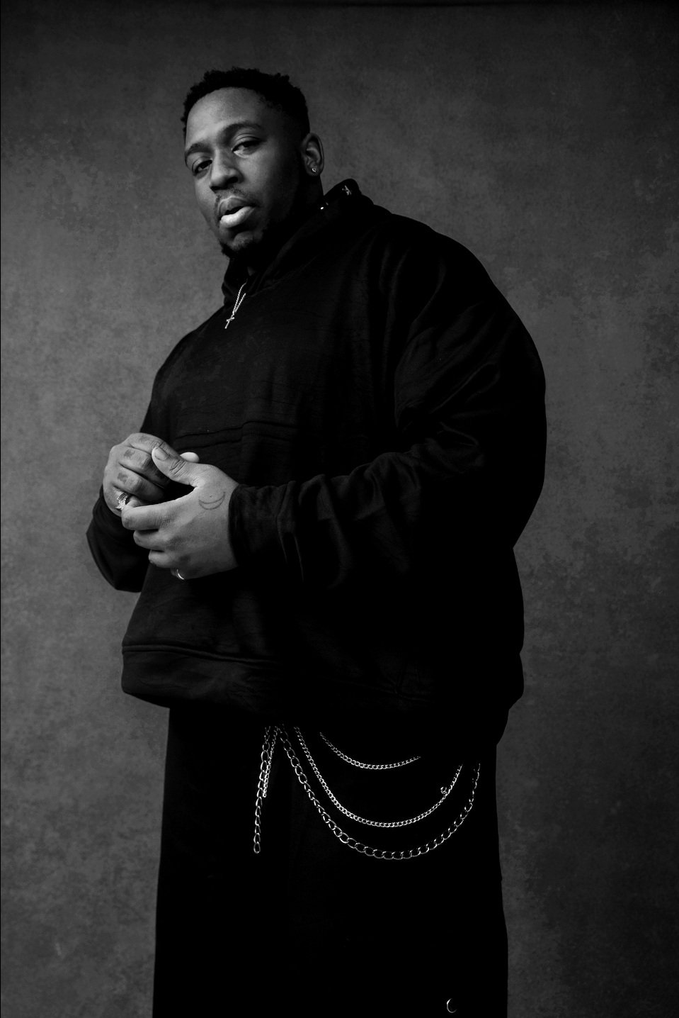 Black and white portrait of a man in a black hoodie, with his hands clasped and chains hanging from his waist, standing against a dark background.