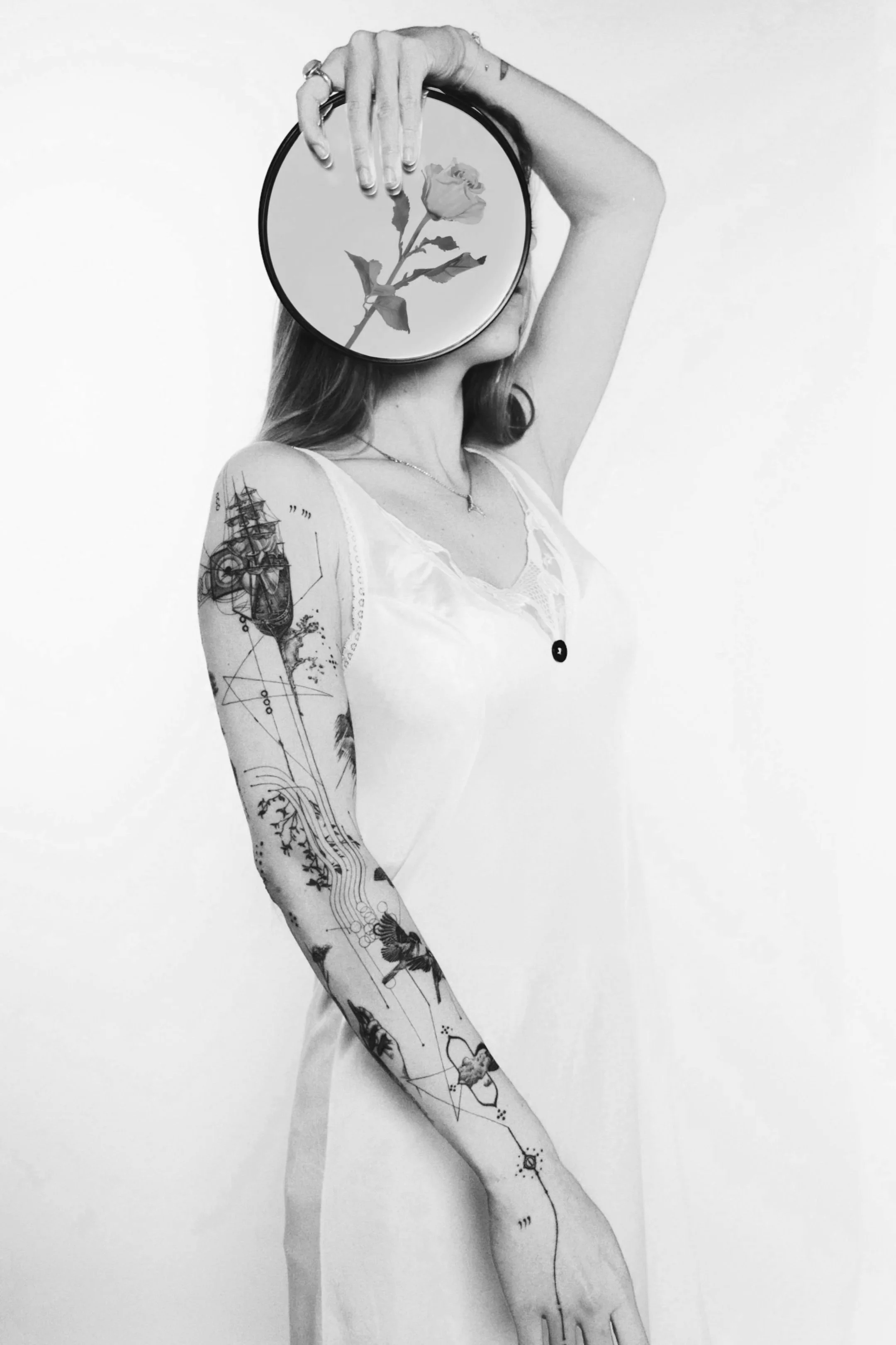 A woman covering her face with a round mirror showing a rose, while her body and arm are covered in tattoos, in black and white.