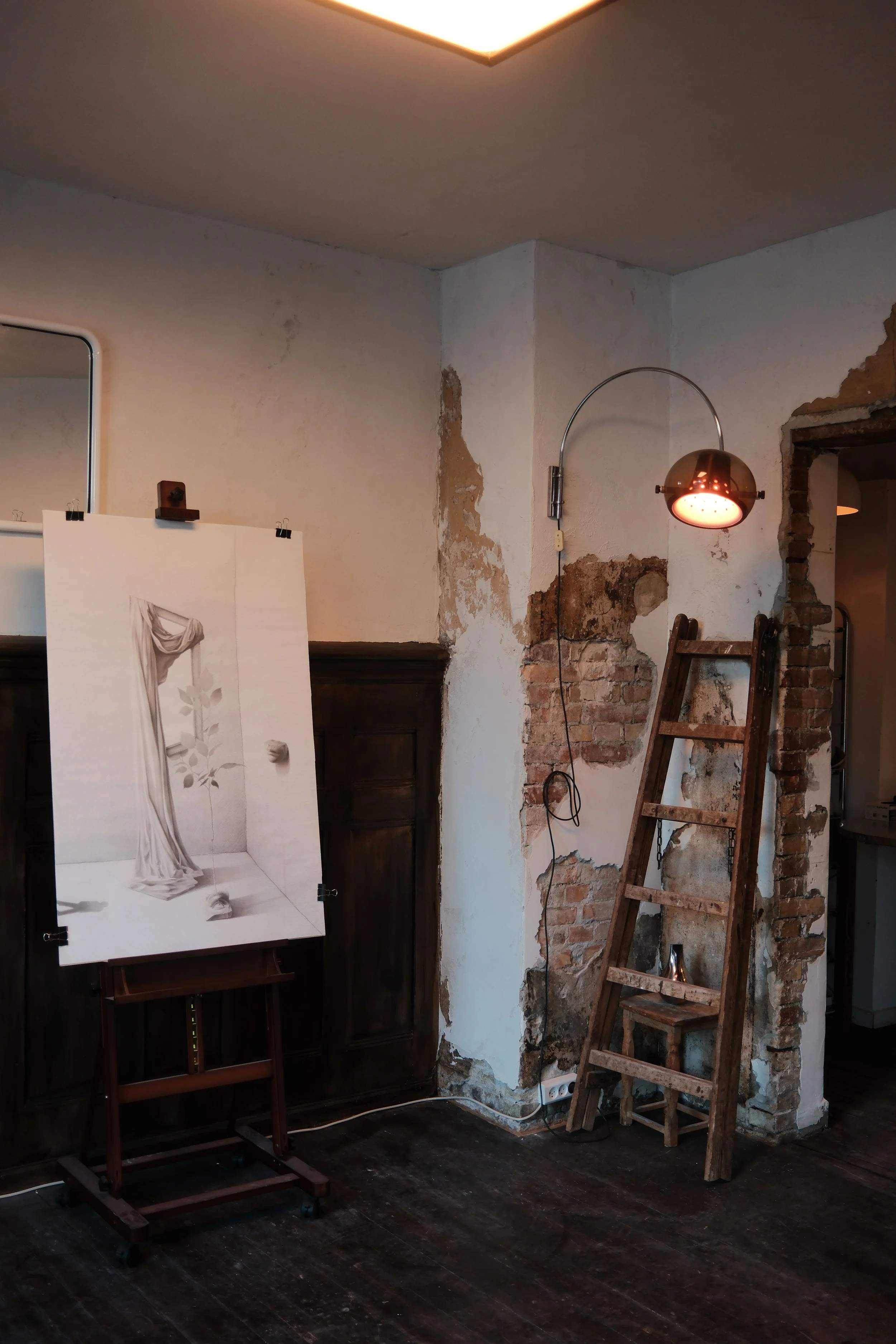An interior space with a damaged wall and exposed brickwork, a wooden ladder, an adjustable wall lamp with a copper finish, a mirror, and a drawing board with a sketch of a draped curtain and flowers.