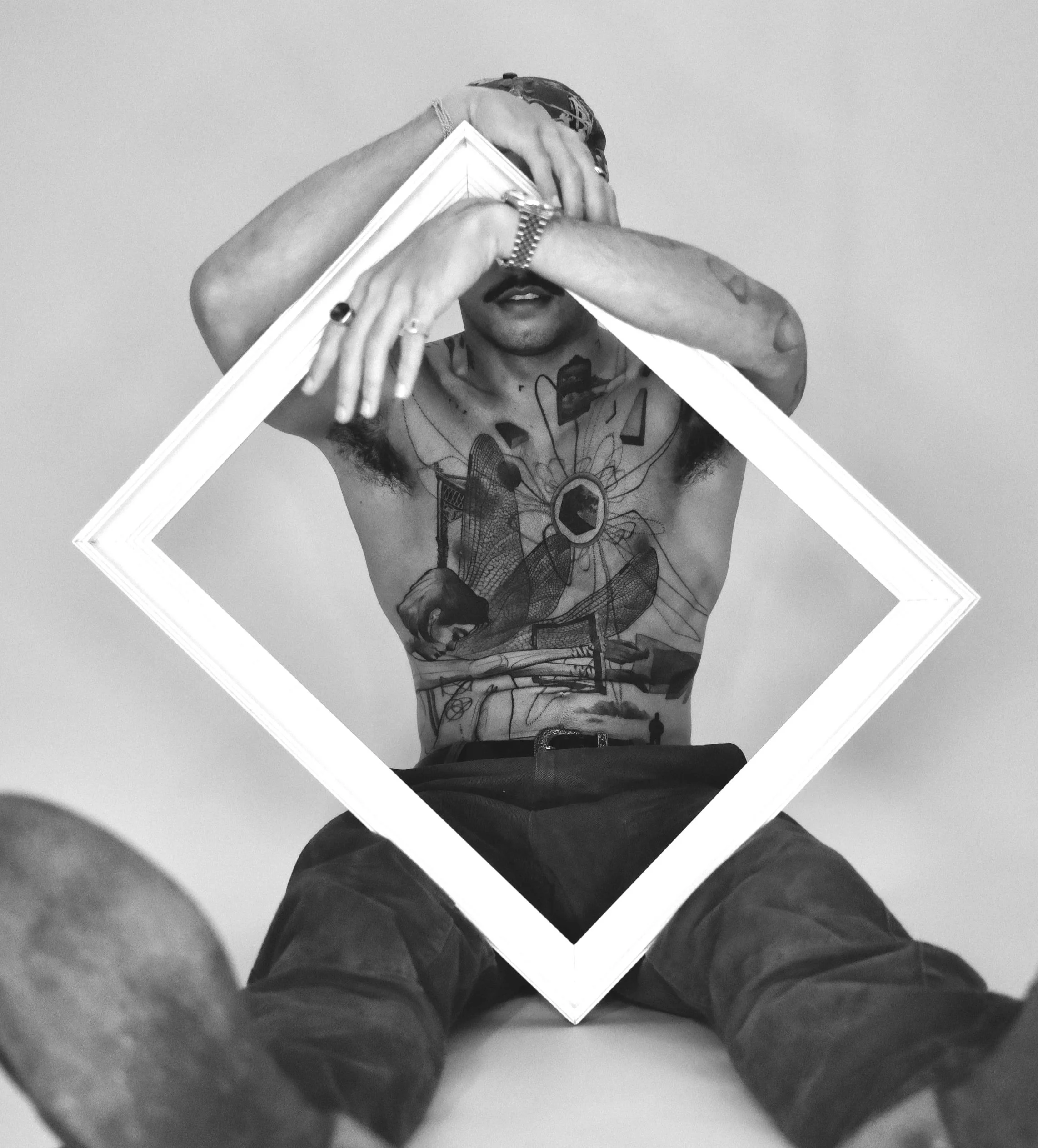 Black and white photo of a person with tattoos on their chest, wearing a bandana, holding a framed empty picture frame over their face, with only part of their mouth visible, taken from a low angle.