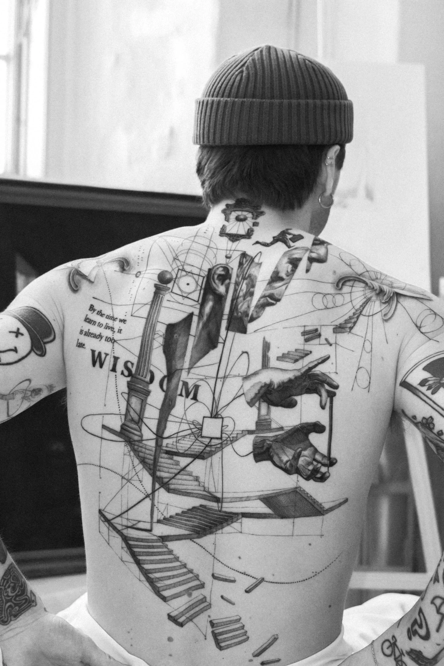 A person with short hair wearing a beanie and earrings, with extensive tattoos on their back and arms. The tattoos include geometric designs, a staircase, and various images.