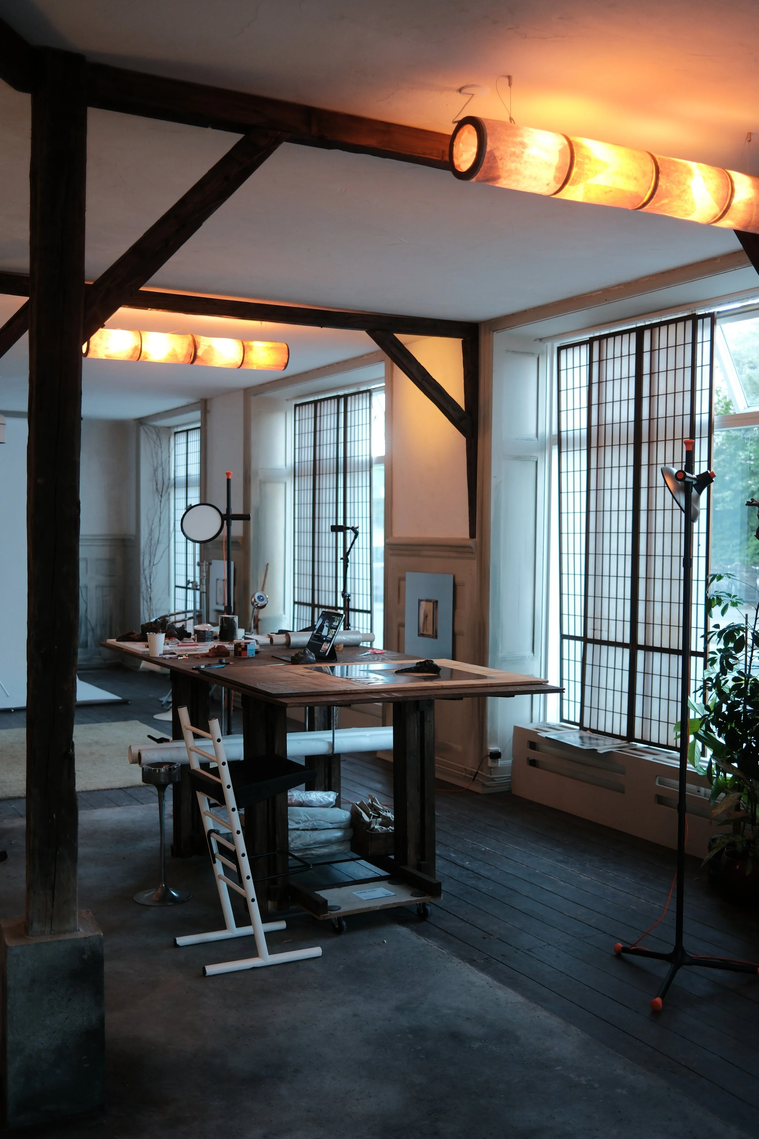 Interior of a room under renovation with large windows, wooden beams, a workstation with tools, and a lighting fixture on a stand.