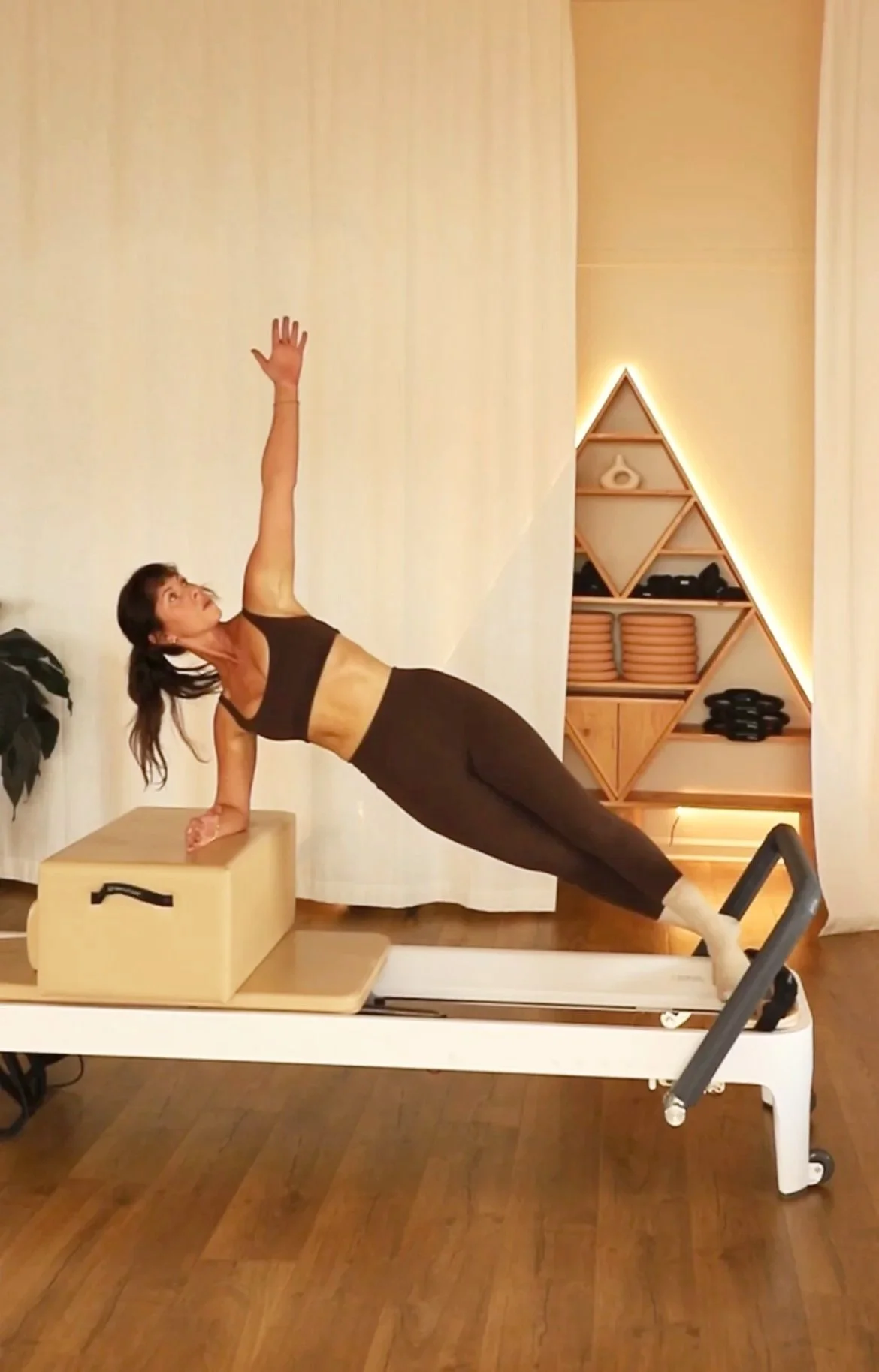 A woman in athletic wear performs a side plank exercise with her hand on a raised platform and her feet on a treadmill, in a room with wooden floors and decorative shelving with lighting.