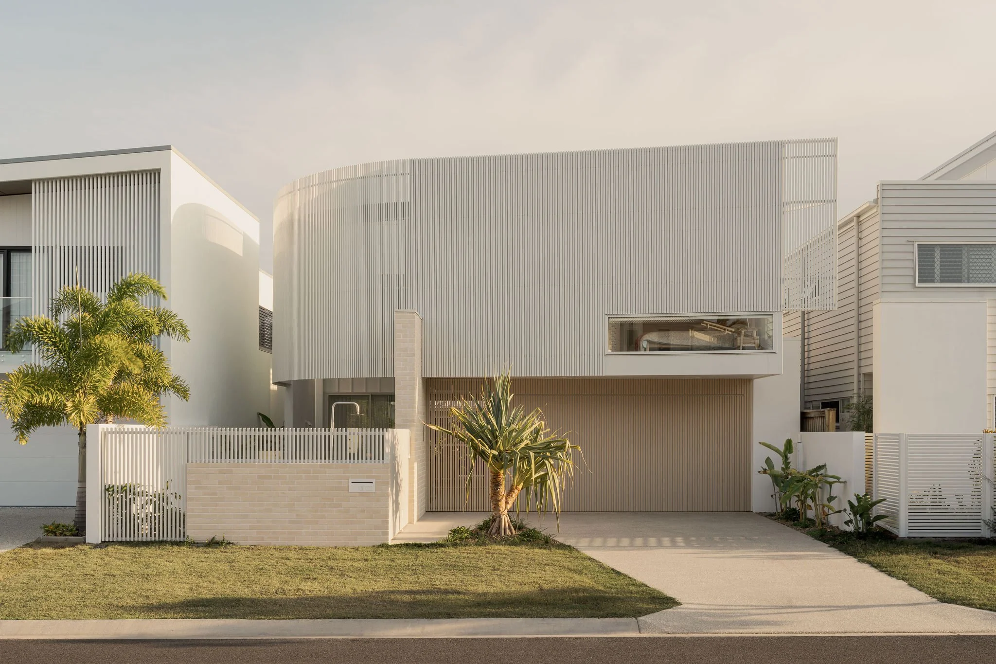 A stunning two-storey home with a curved white facade, brick fencing, and a landscaped front yard