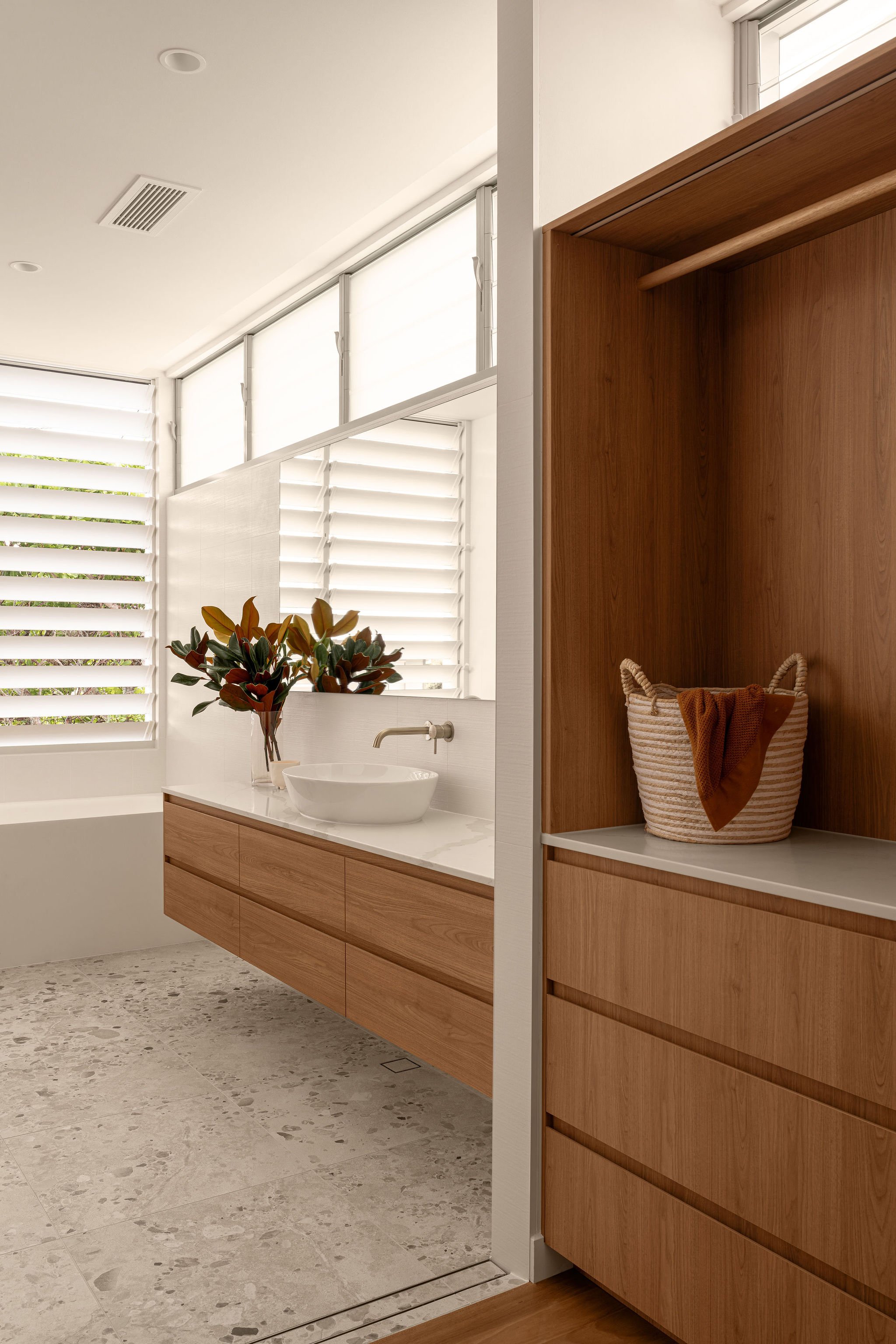 Detail of a modern ensuite vanity with timber drawers, a vessel basin, and brushed nickel tapware, positioned beside a woven basket nook and under a large horizontal window with louvre shutters