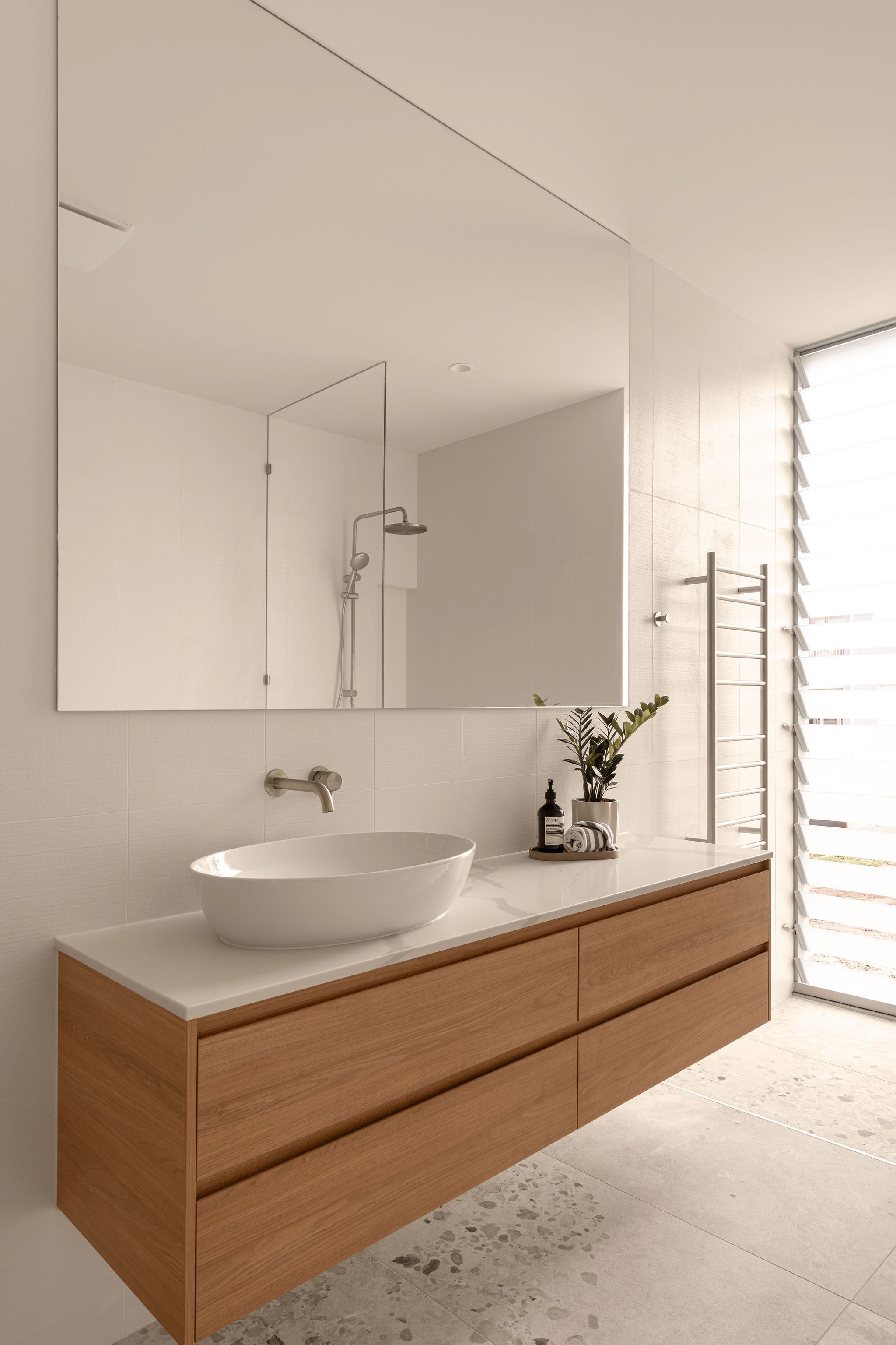 Minimalist bathroom vanity with a white benchtop, timber drawers, and vessel basin, paired with a large mirror and brushed nickel wall tapware, adjacent to a glass-walled shower