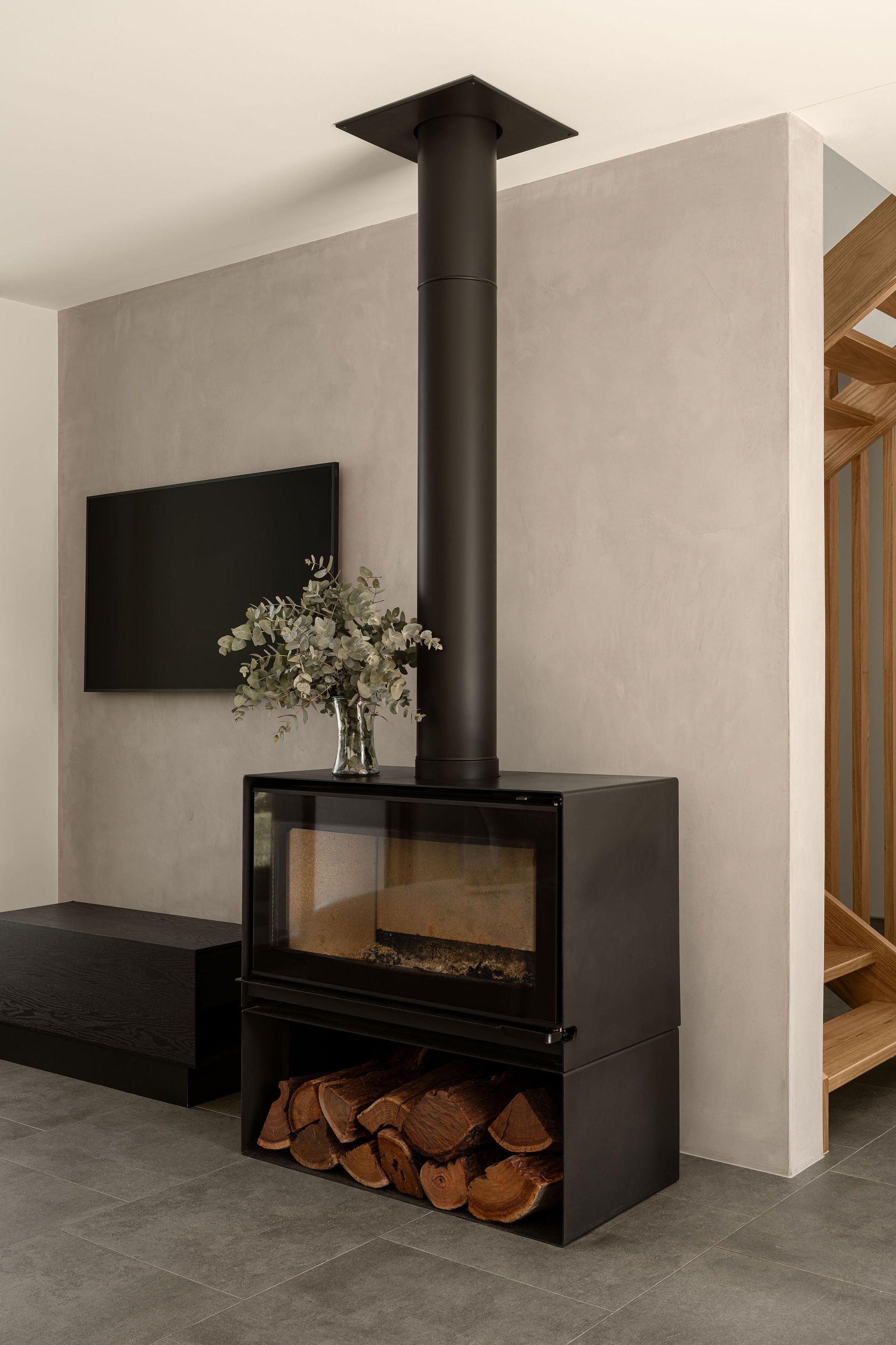 Modern black freestanding fireplace with timber storage underneath, set against a smooth rendered wall, complemented by a TV and a staircase with timber balustrades