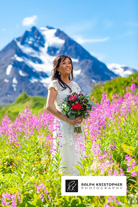  A bride holding red flowers with a mountain in background with fireweed flowers around her and photo by Ralph Krisopher Photography 