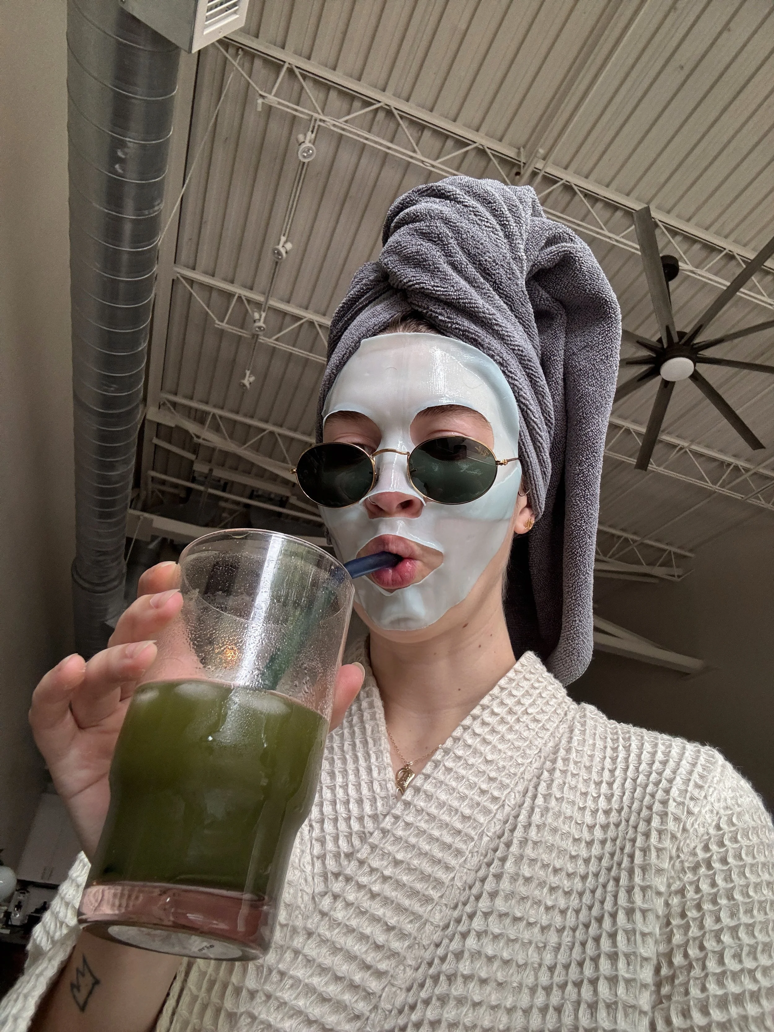 Woman in bathrobe and face mask sipping green juice: morning self-care routine, relaxation at home.
