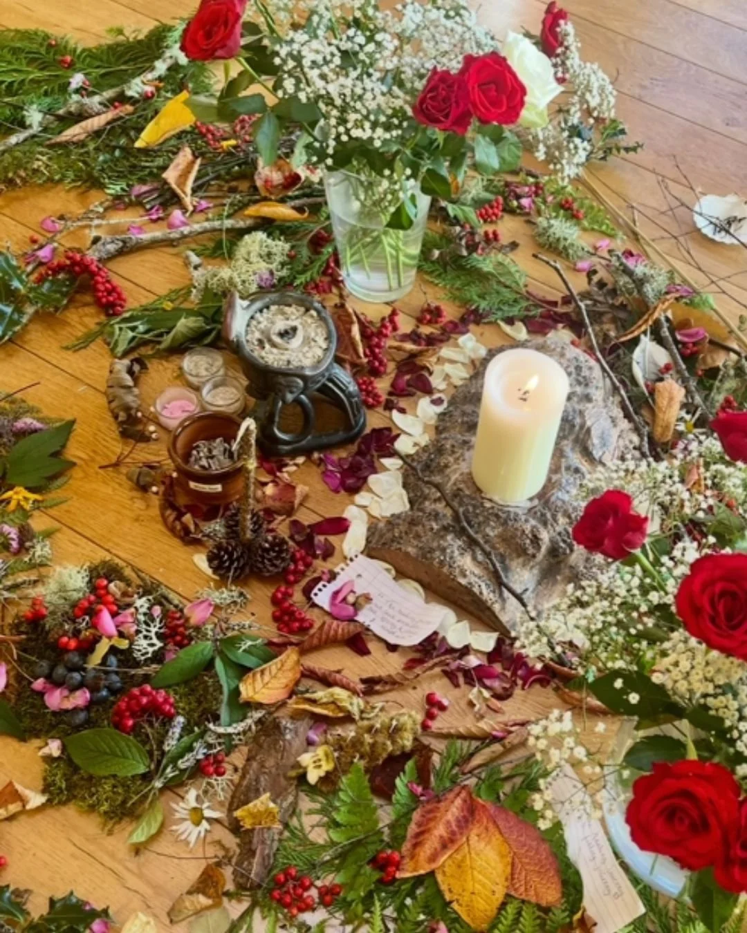 A ceremony offerings, Simone was involved in. An arrangement on a wooden floor featuring roses, baby’s breath, leaves, pinecones, candles, and notes.