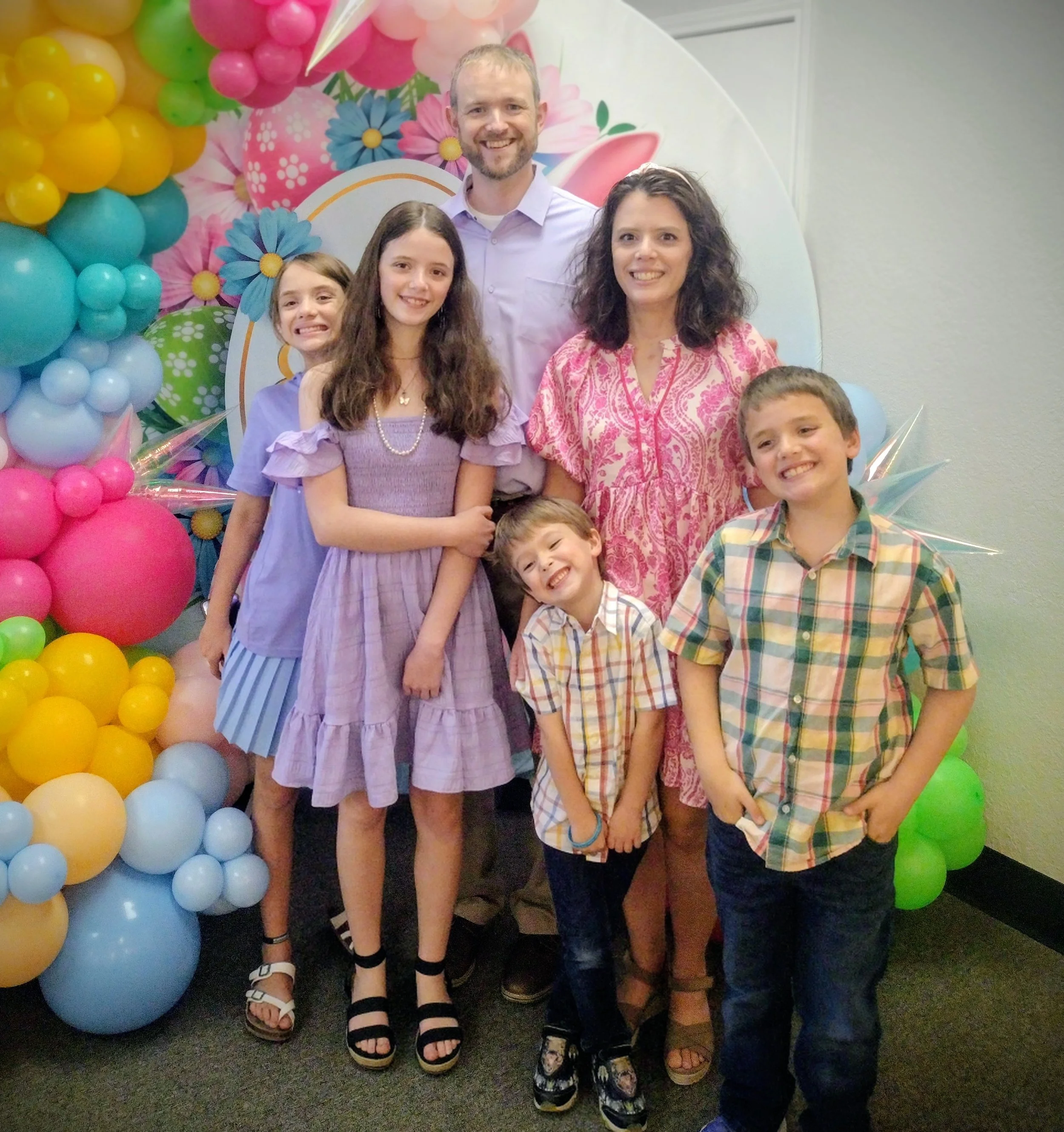 A group of seven people, including three adults and four children, smiling and posing in front of a colorful balloon backdrop at a celebration.