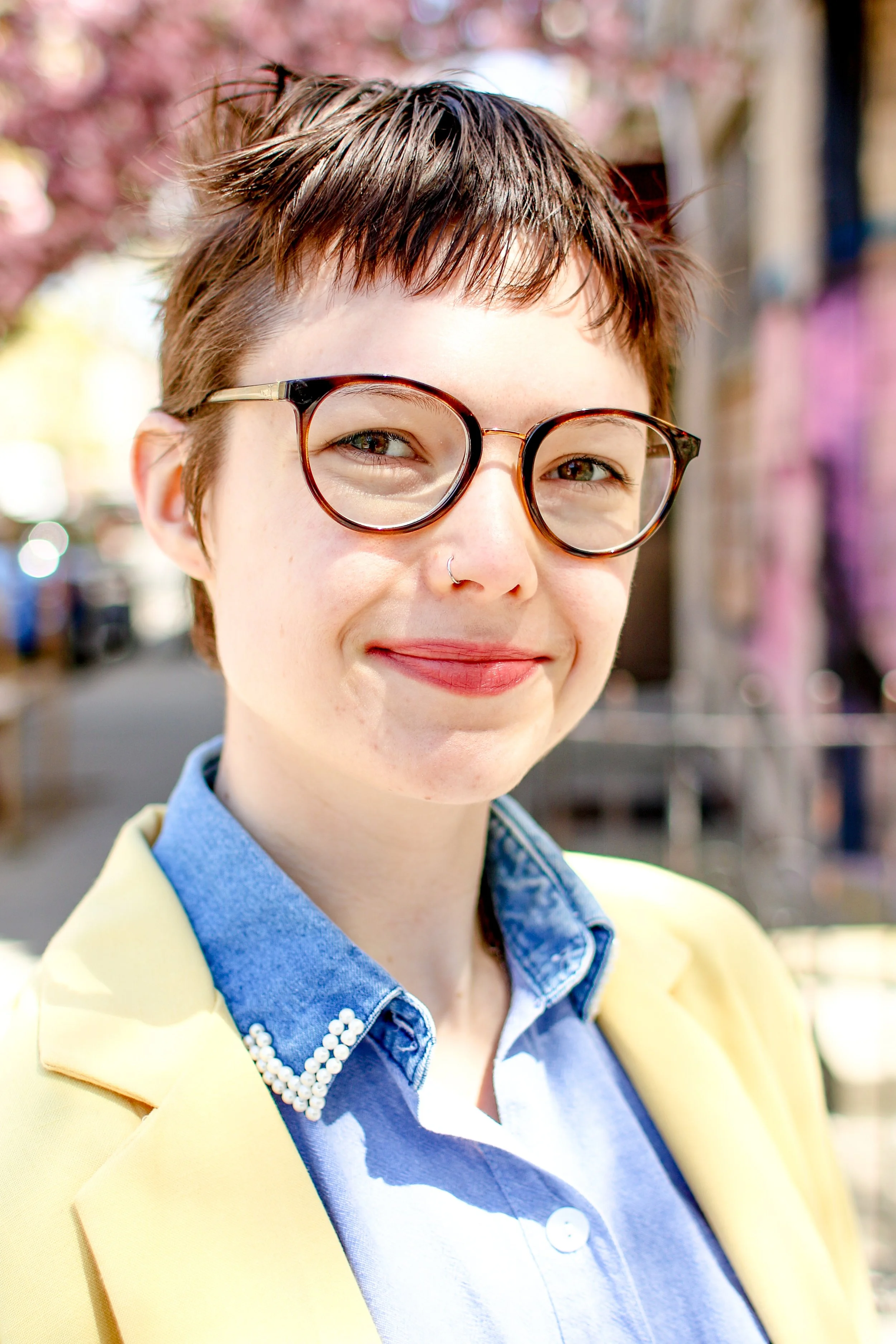 A brown haired person with round glasses wearing a yellow blazer and blue button-up smiles at the camera in front of purple flowers.