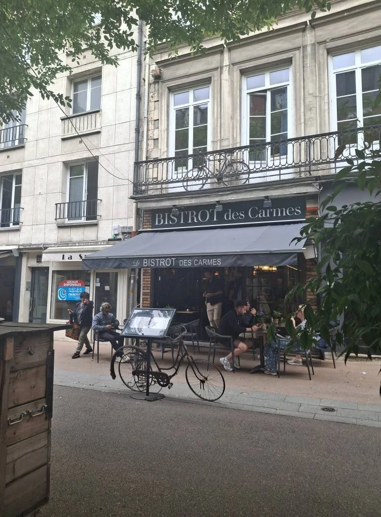 A street view featuring a restaurant named BISTROT des Carmes with outdoor seating, a decorative bicycle sculpture in front with trees and a multi-story building in the background. Des Carmes from Death 2 My Past.