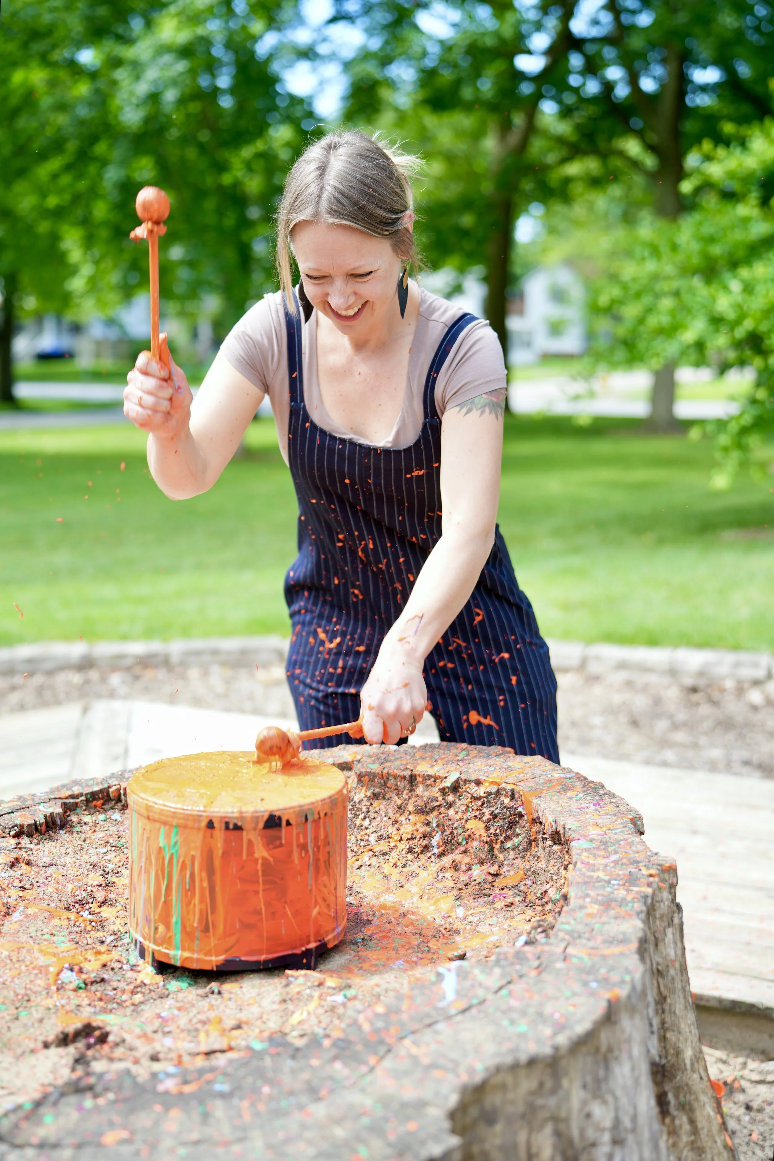 A woman standing outdoors next to two decorated trash cans, smiling and gesturing towards the cans, in front of a brick building with windows that spell out 'CREATE'.