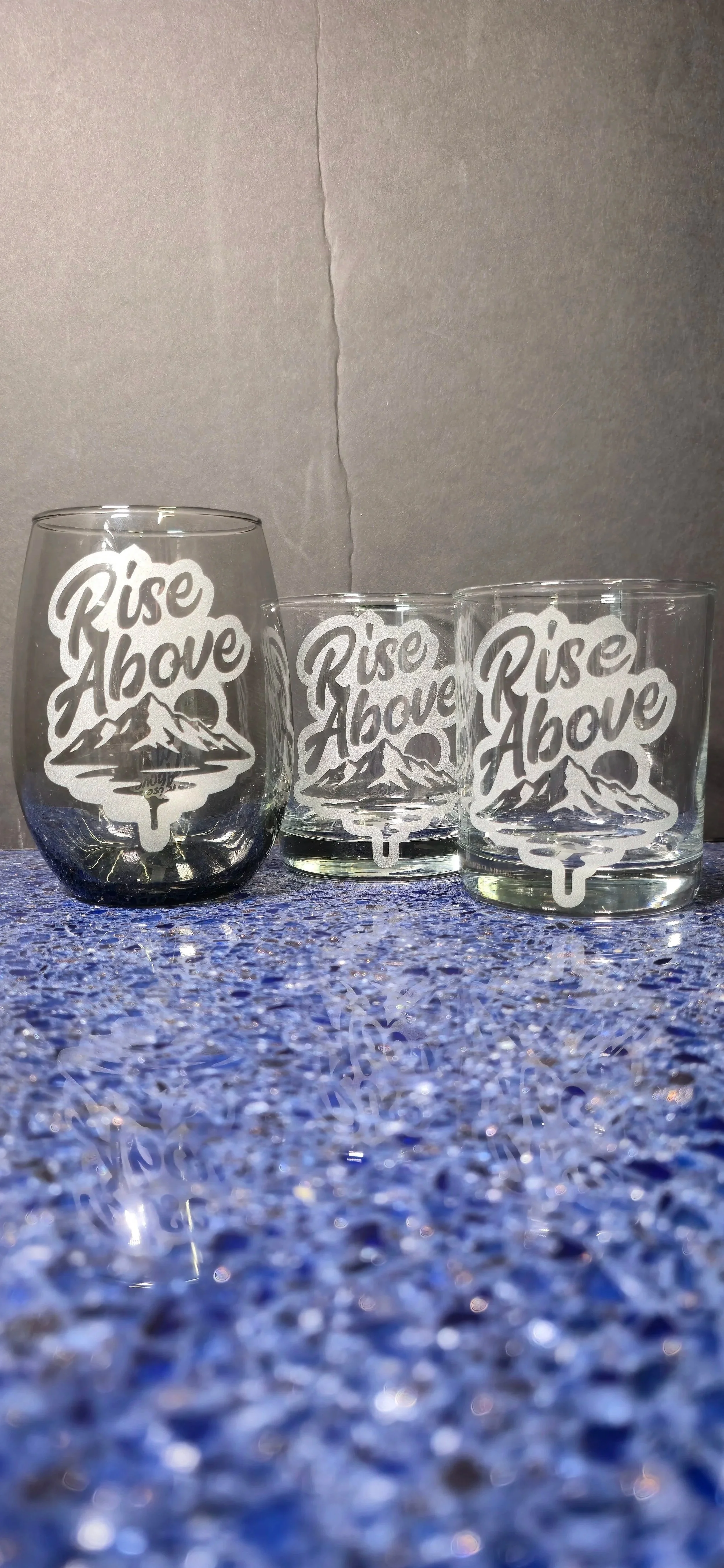 Three glasses with the phrase 'Rise Above' and a mountain design printed on them, placed on a blue textured surface.