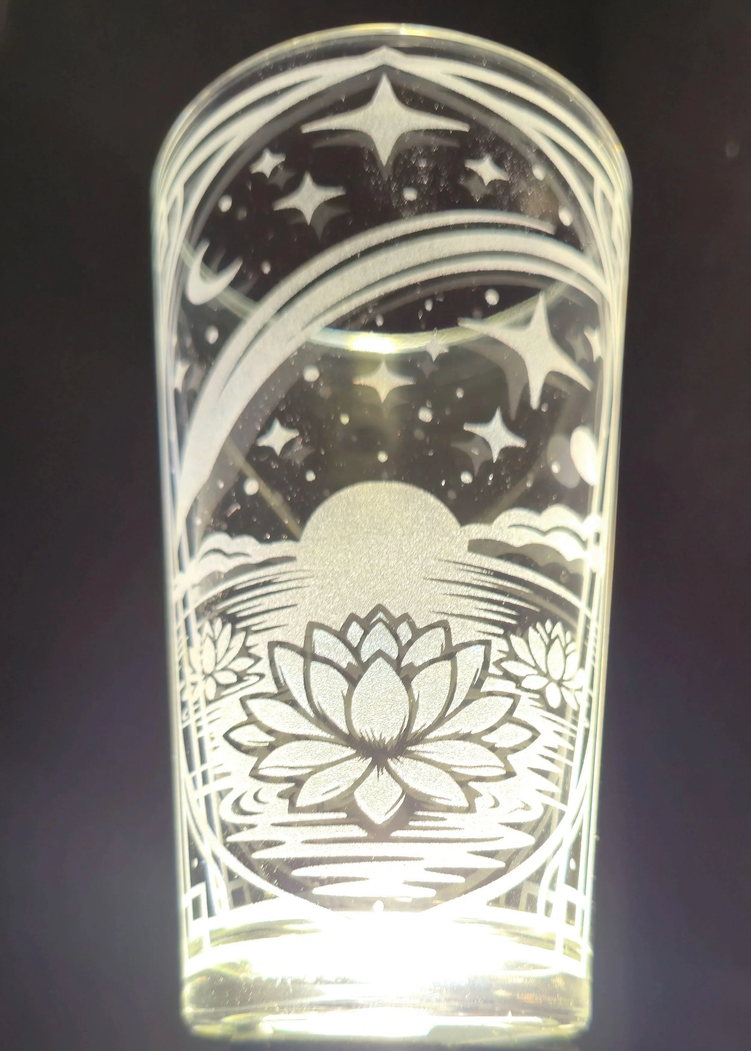 A glass decorative piece featuring an engraved design of a lotus flower, a full moon, stars, and water reflections, with a dark background.