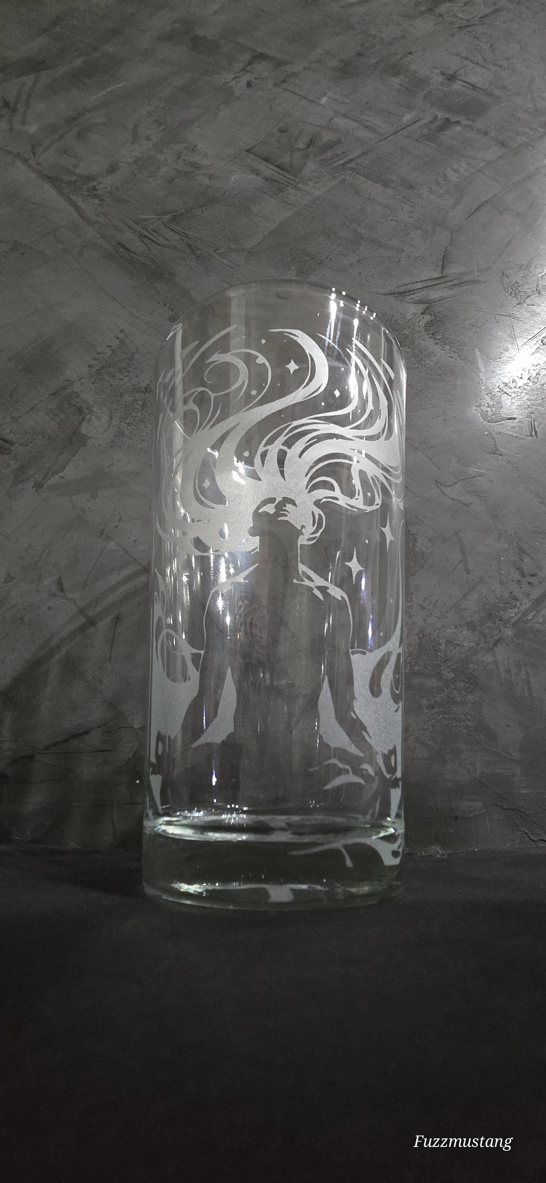 A glass with an etching of a mythical creature with flowing hair and a crown, surrounded by stars and swirling patterns, placed on a dark surface against a textured gray wall.
