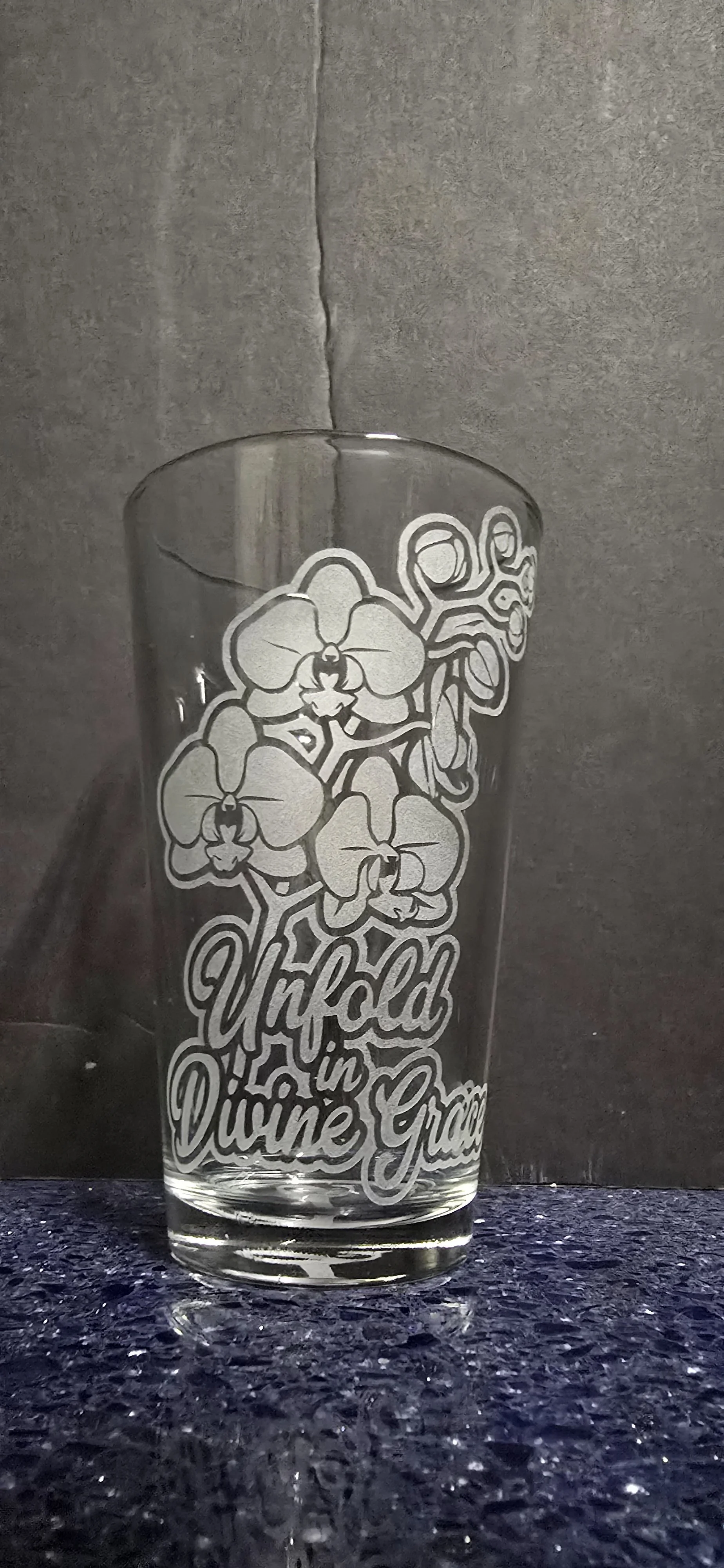Clear glass with a white etching of orchids and the text "Unfold in Divine Grace" standing on a dark textured surface against a gray background.