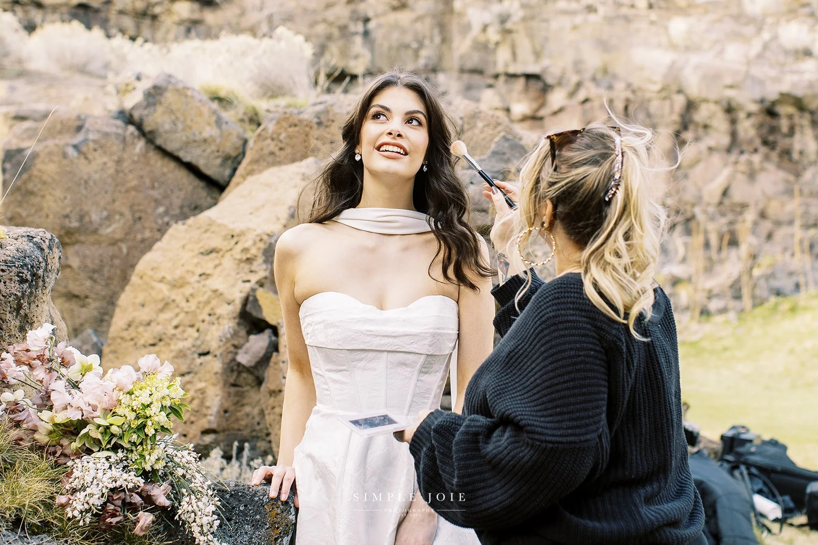 A woman in a white strapless dress with a turtleneck collar is having makeup applied outdoors in front of rocks, with a bouquet of flowers nearby.