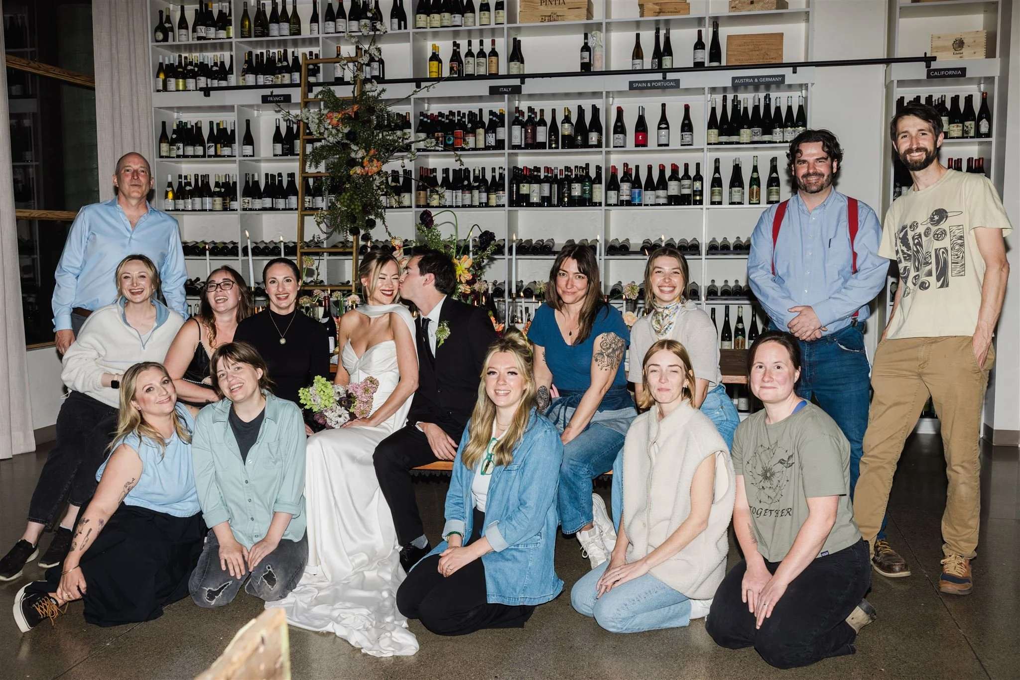 A group of people celebrating a wedding, with the bride and groom seated in the center, surrounded by friends and family in a wine cellar setting with wine bottles on shelves behind them.
