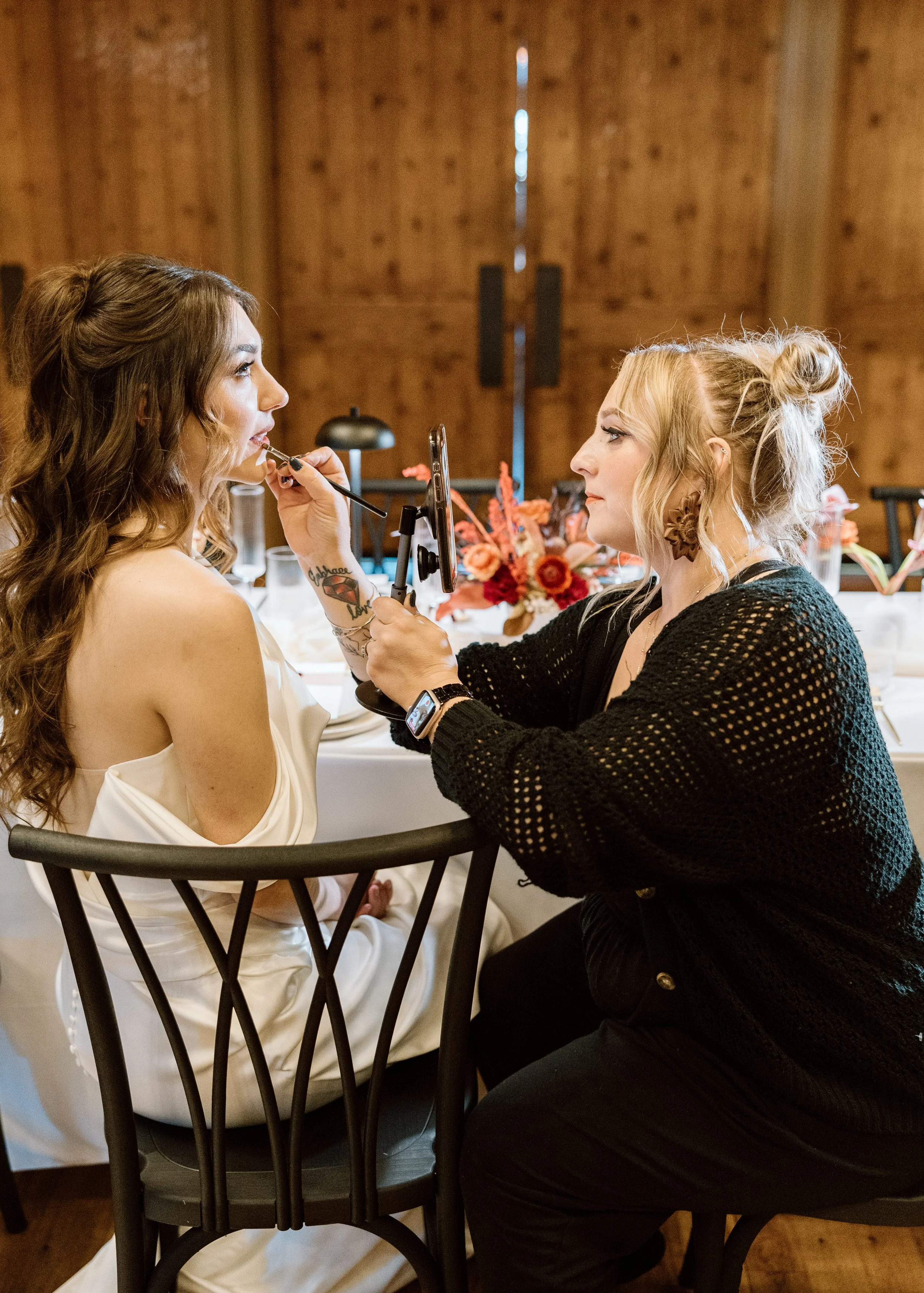A woman with long curly brown hair getting her makeup done by a makeup artist with blonde hair in an updo, in a rustic wooden venue with a table decorated with flowers in the background.