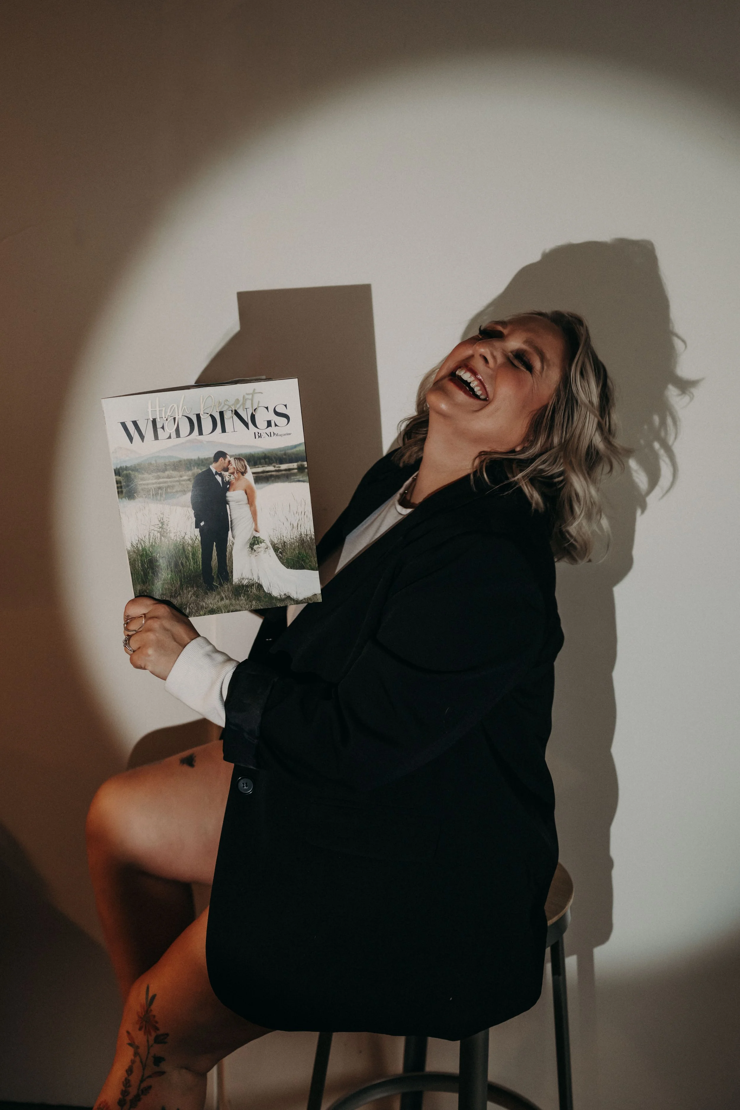 A woman with short, curly blonde hair, dressed in a black blazer, sitting on a stool, holding a wedding magazine, and laughing joyfully. She has tattoos on her left leg, and the photo captures her casting a shadow on the wall behind her.