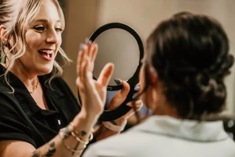 A makeup artist shows a woman her reflection in a small hand mirror during a makeup session.
