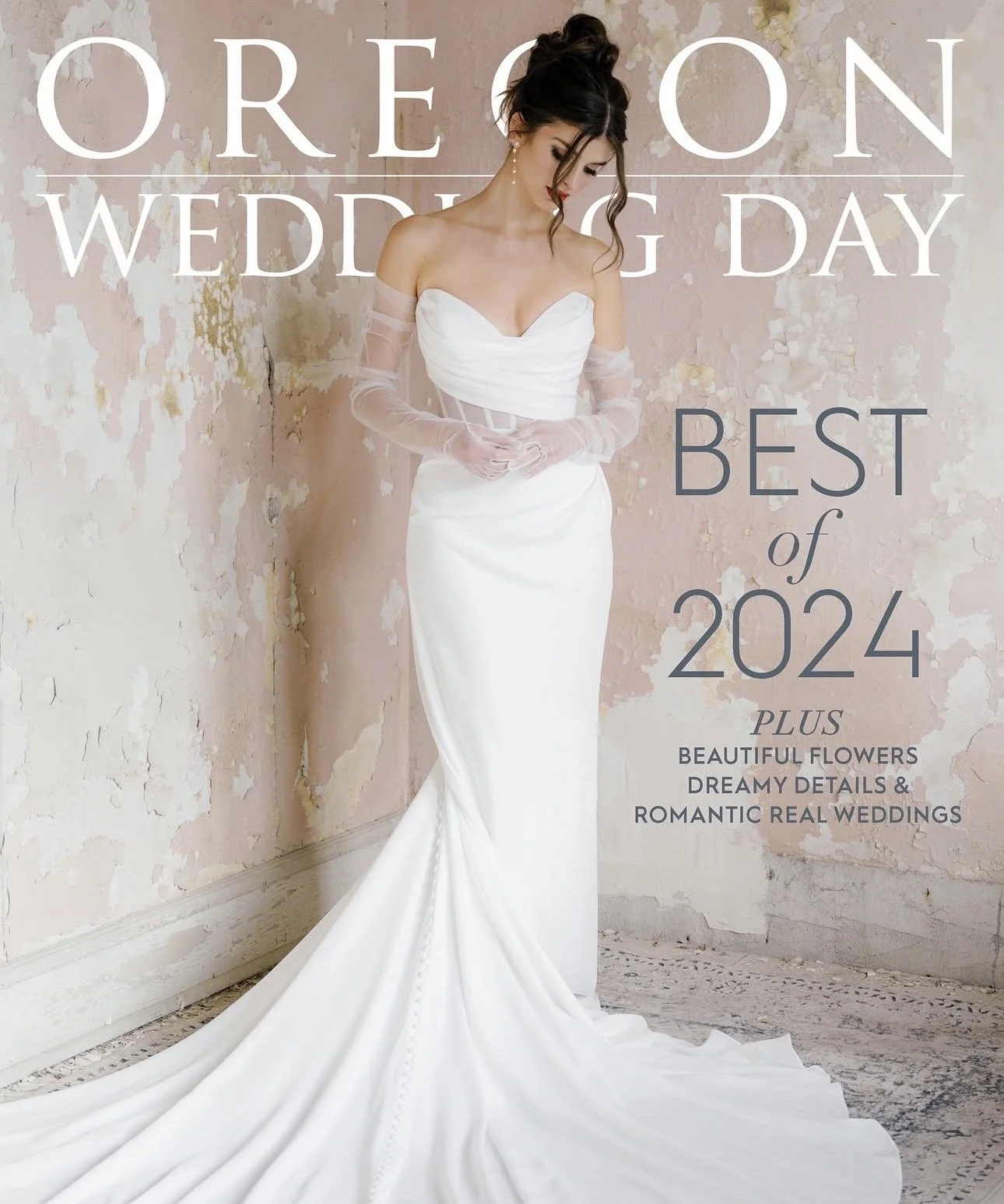 A woman in a white wedding dress with sheer sleeves and a sweetheart neckline, standing against a weathered, pinkish wall.