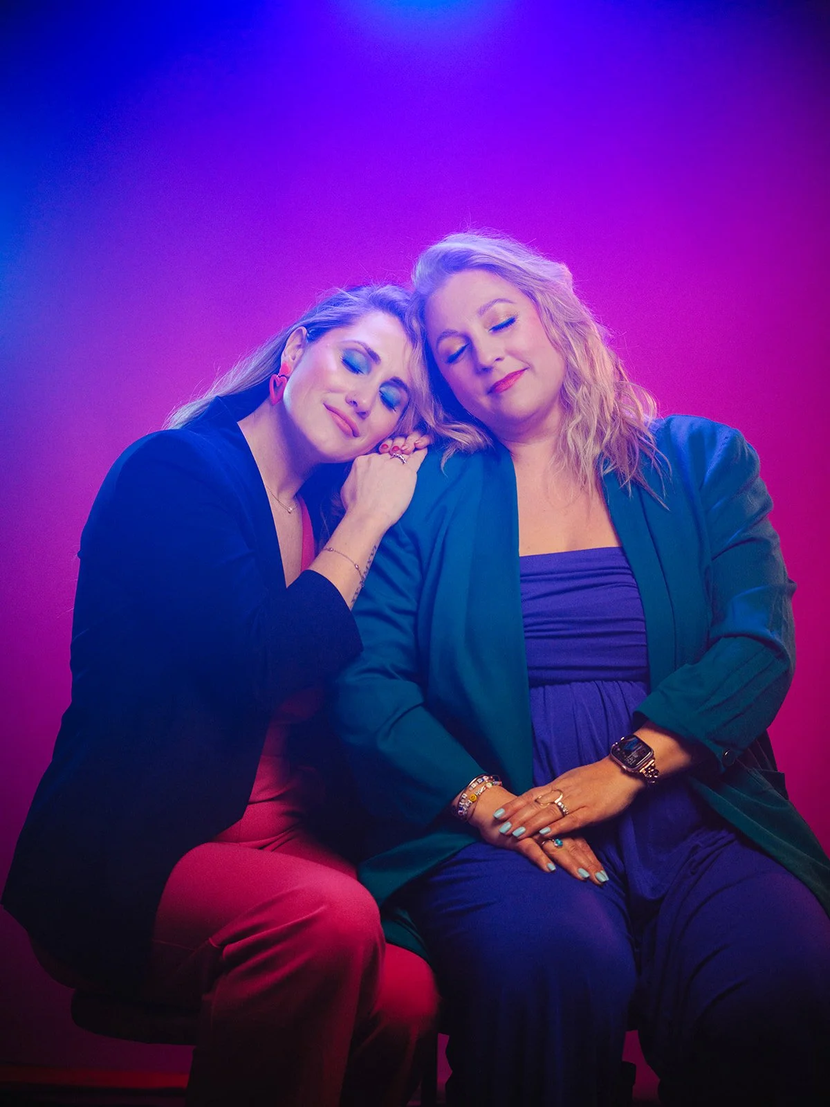 Two women sitting close together under purple and pink lighting, with their eyes closed and peaceful expressions.