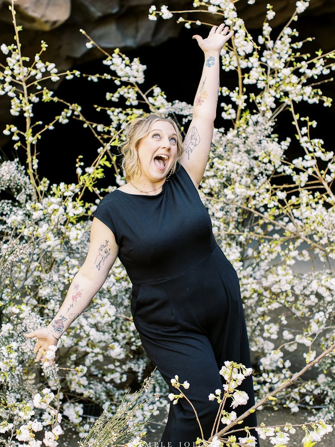 Woman in black outfit smiling and posing with arm raised among blooming branches of white flowers.