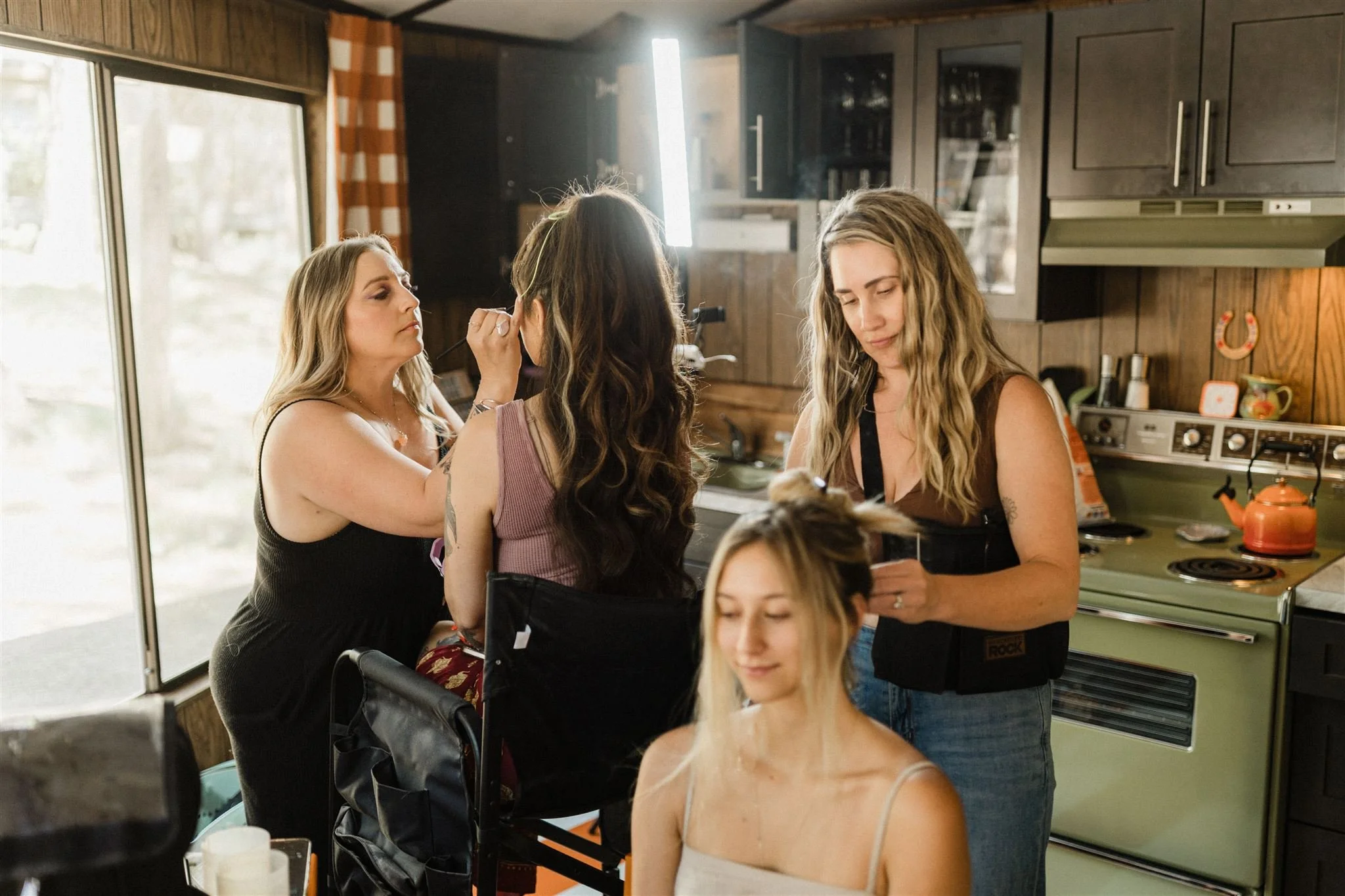 Women preparing for a photo shoot in a kitchen, with one woman applying makeup and another adjusting hair, while a model sits with her eyes closed and smiling.
