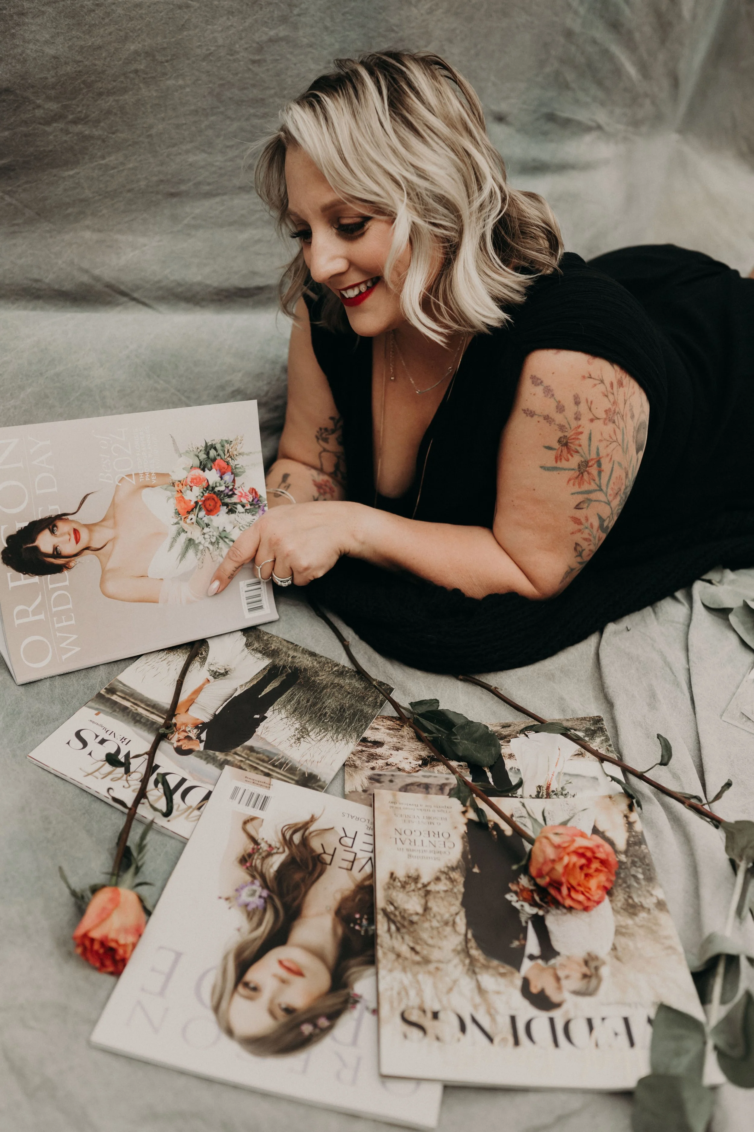 A woman with blonde wavy hair and tattoos, lying on a gray surface, looking at wedding magazines surrounded by flowers and greenery.