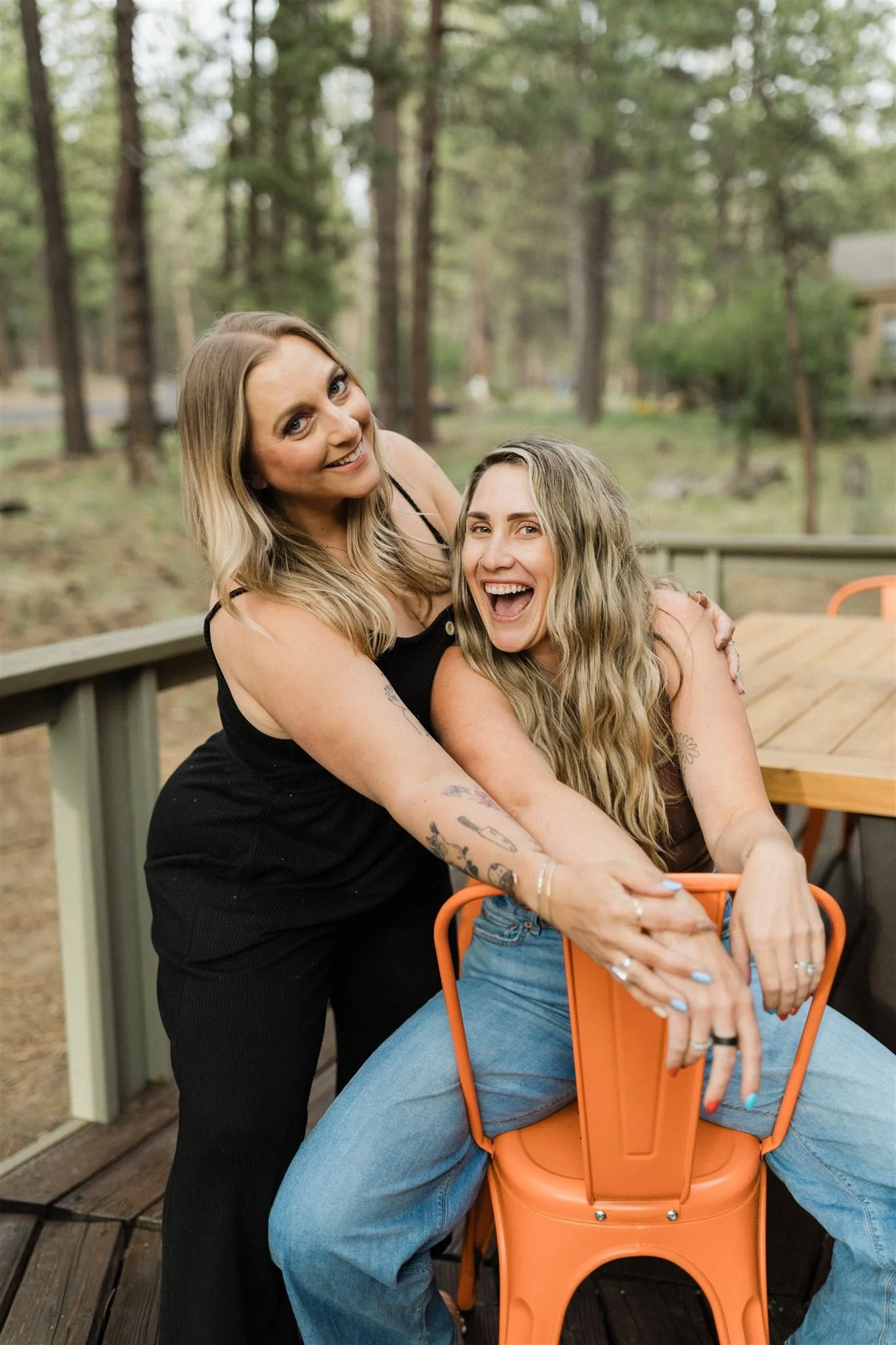Two women, one standing and one sitting, embracing and smiling on a wooden deck outdoors with a forest in the background.