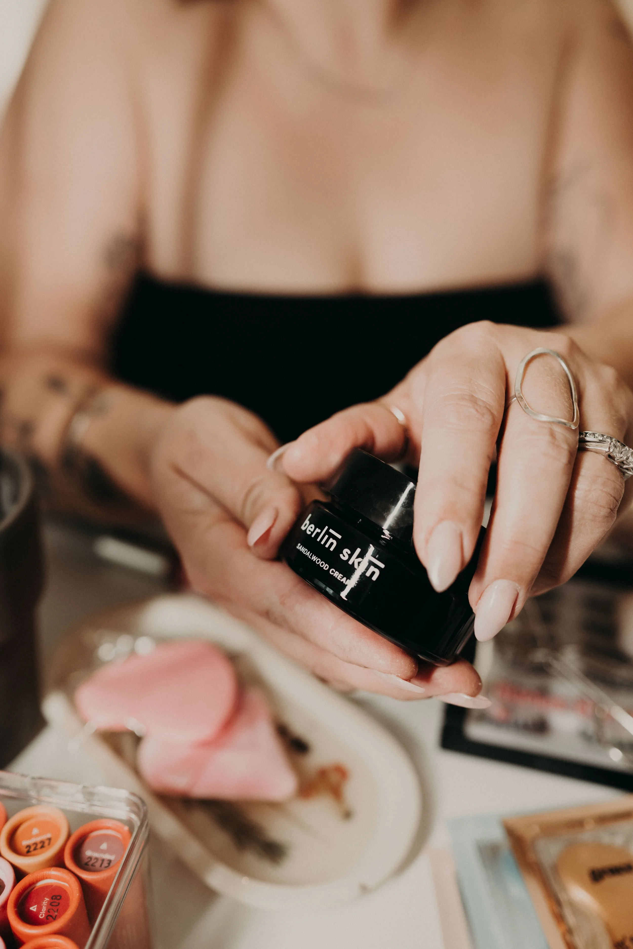 A woman with tattoos and rings on her fingers holding a small black container of Berlin Skin sandalwood cream with a blurred plate of food and a box of lipsticks in the background.