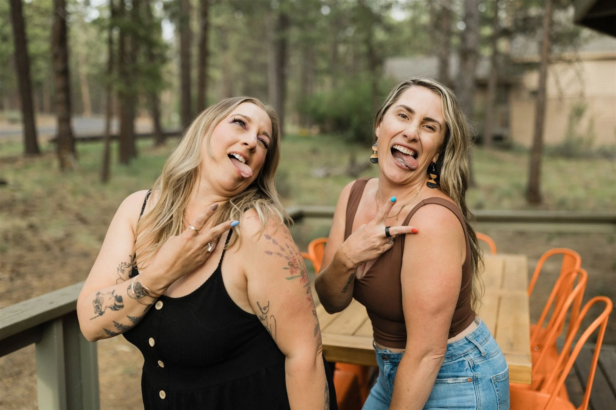 Two women making silly faces and pointing at each other outdoors in a wooded area, with a picnic table and orange chairs in the background.