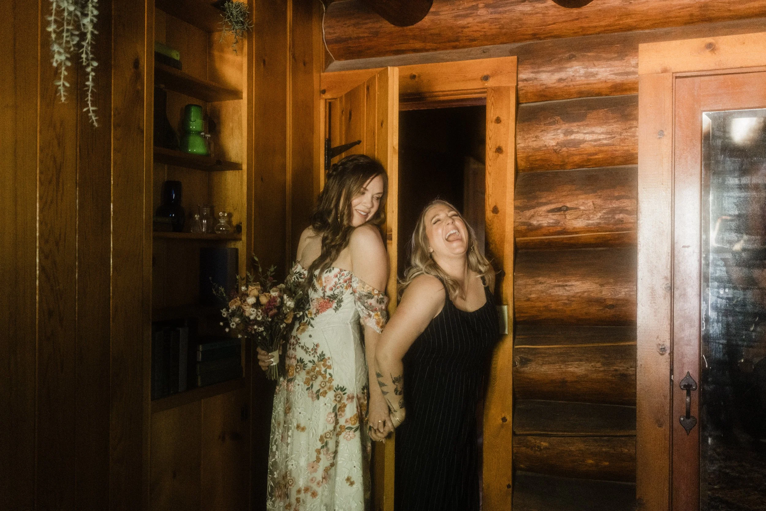 Two women standing in a door frame, holding hands and laughing. One woman is holding a bouquet of flowers and wearing a floral dress, while the other is wearing a black striped dress.