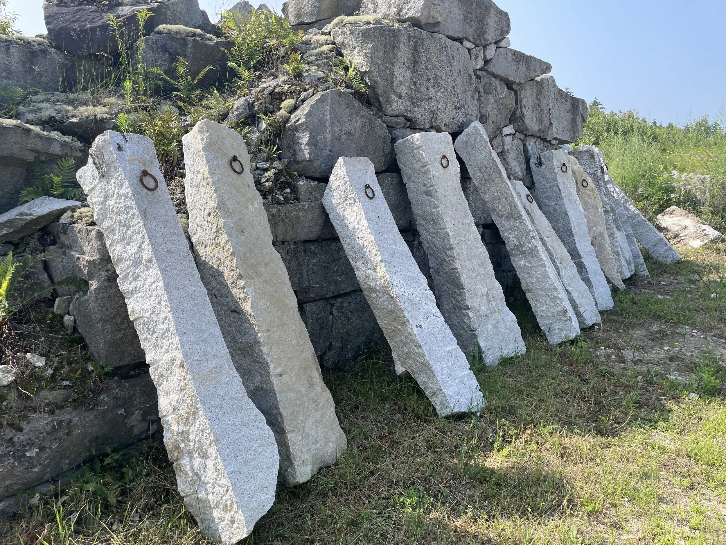 Several large, rough-cut hitching posts with metal rings leaning against a stone wall outdoors on a grassy area.
