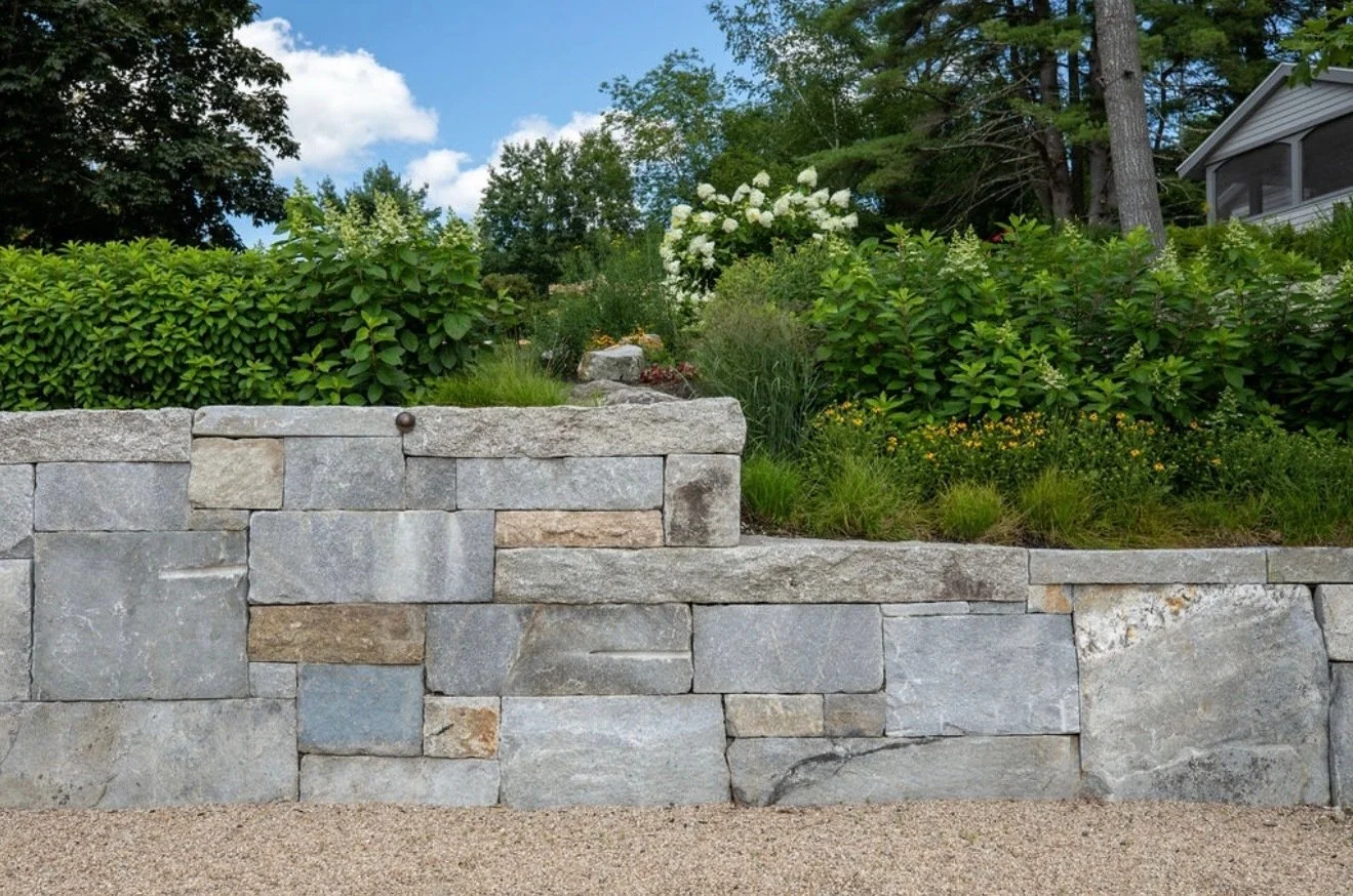 Stone retaining wall with a garden and house in the background, trees, shrubs, and a partly cloudy sky.