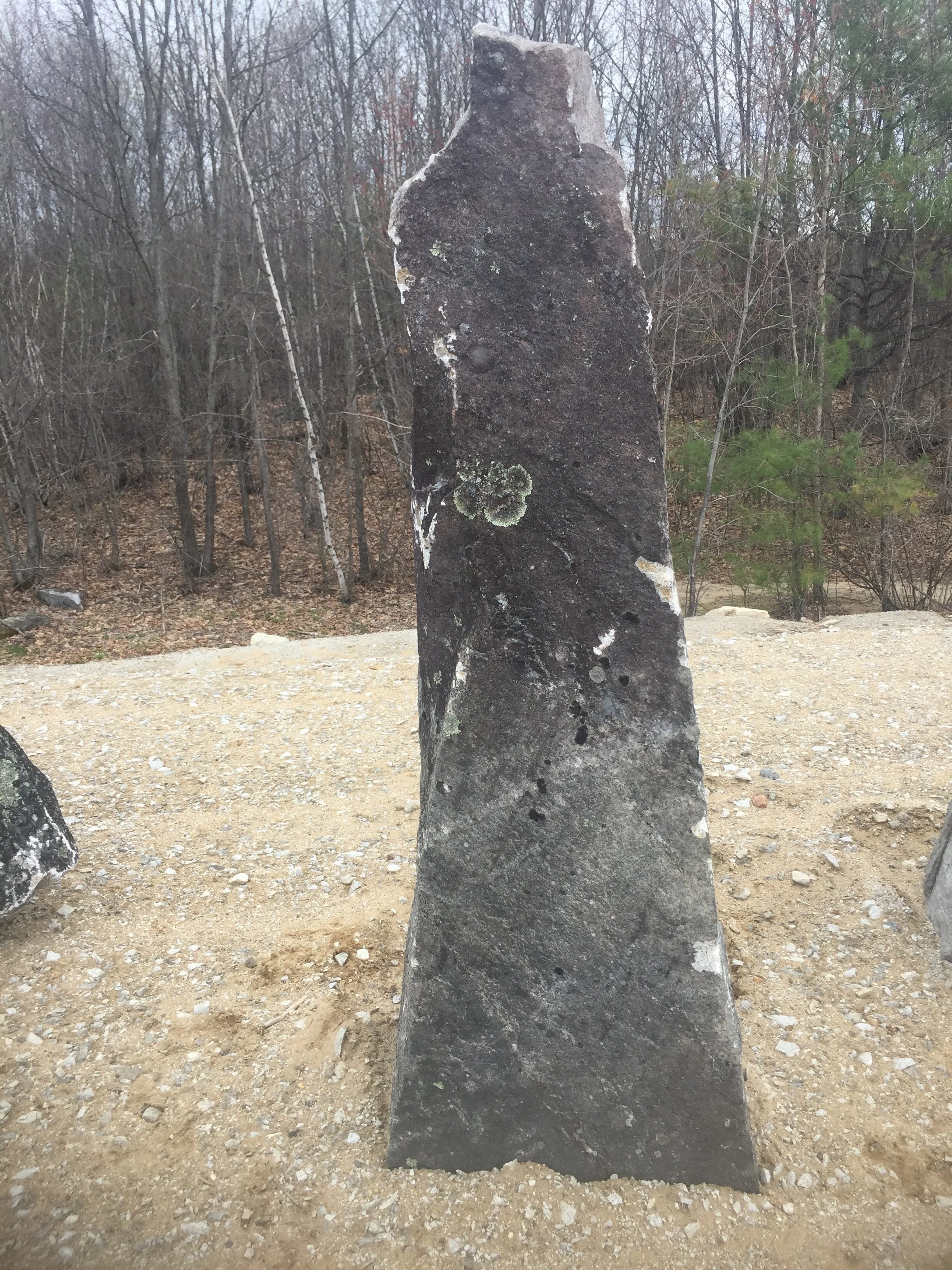 A tall, weathered stone monument standing alone on a gravel surface outdoors with trees in the background.