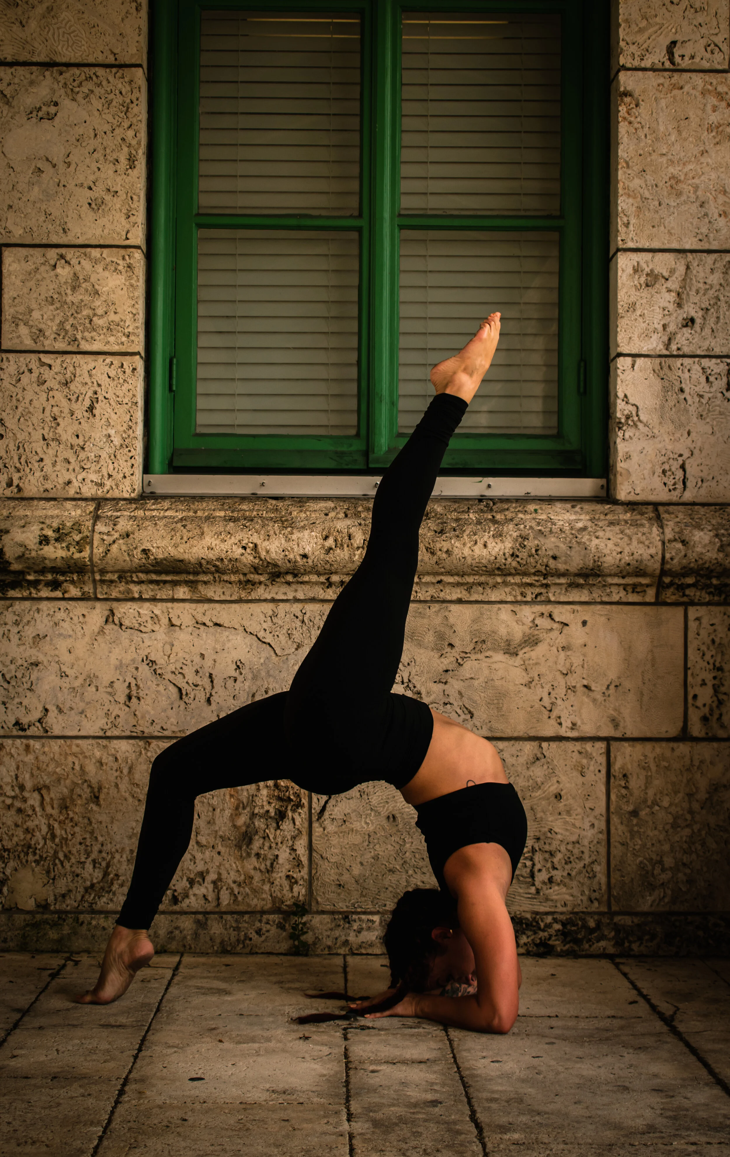 Fitness content shoot with instructor Lexi Ferra holding a one-legged backbend yoga pose against a stone wall with green window frame.