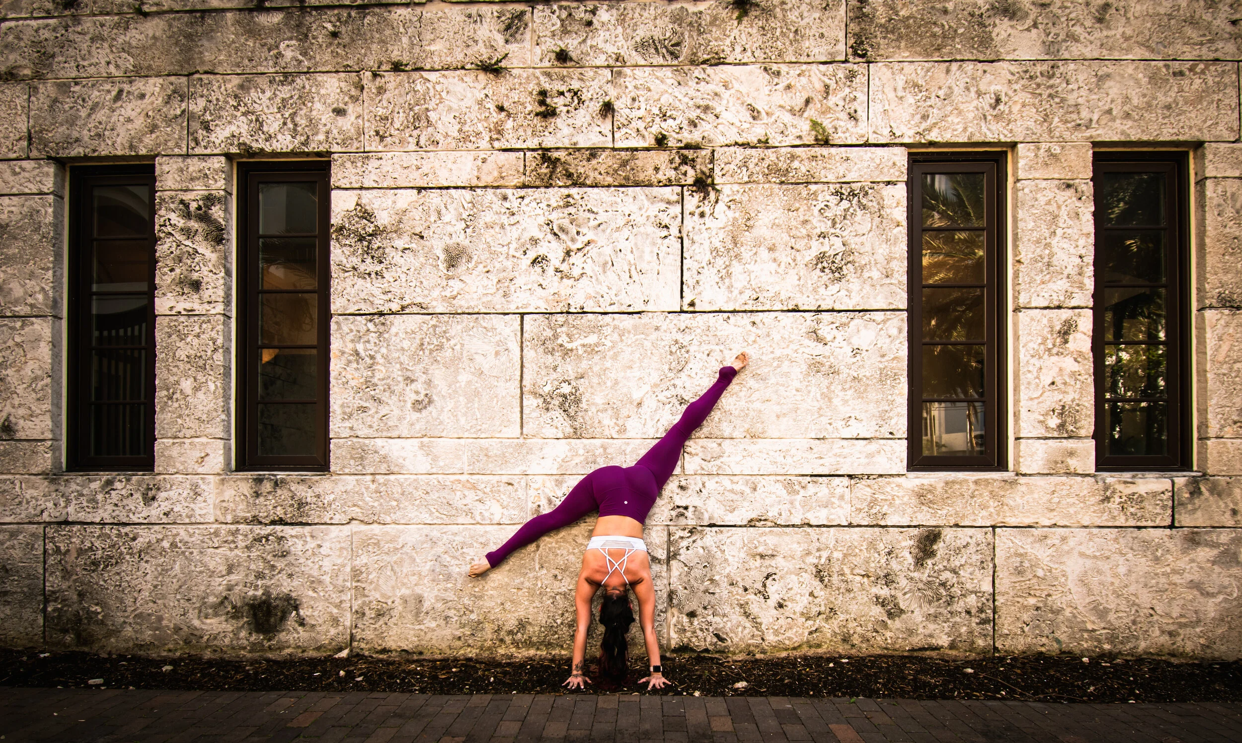 Lexi Ferra holding a freestanding handstand split, framed by a textured stone wall in Coral Gables.