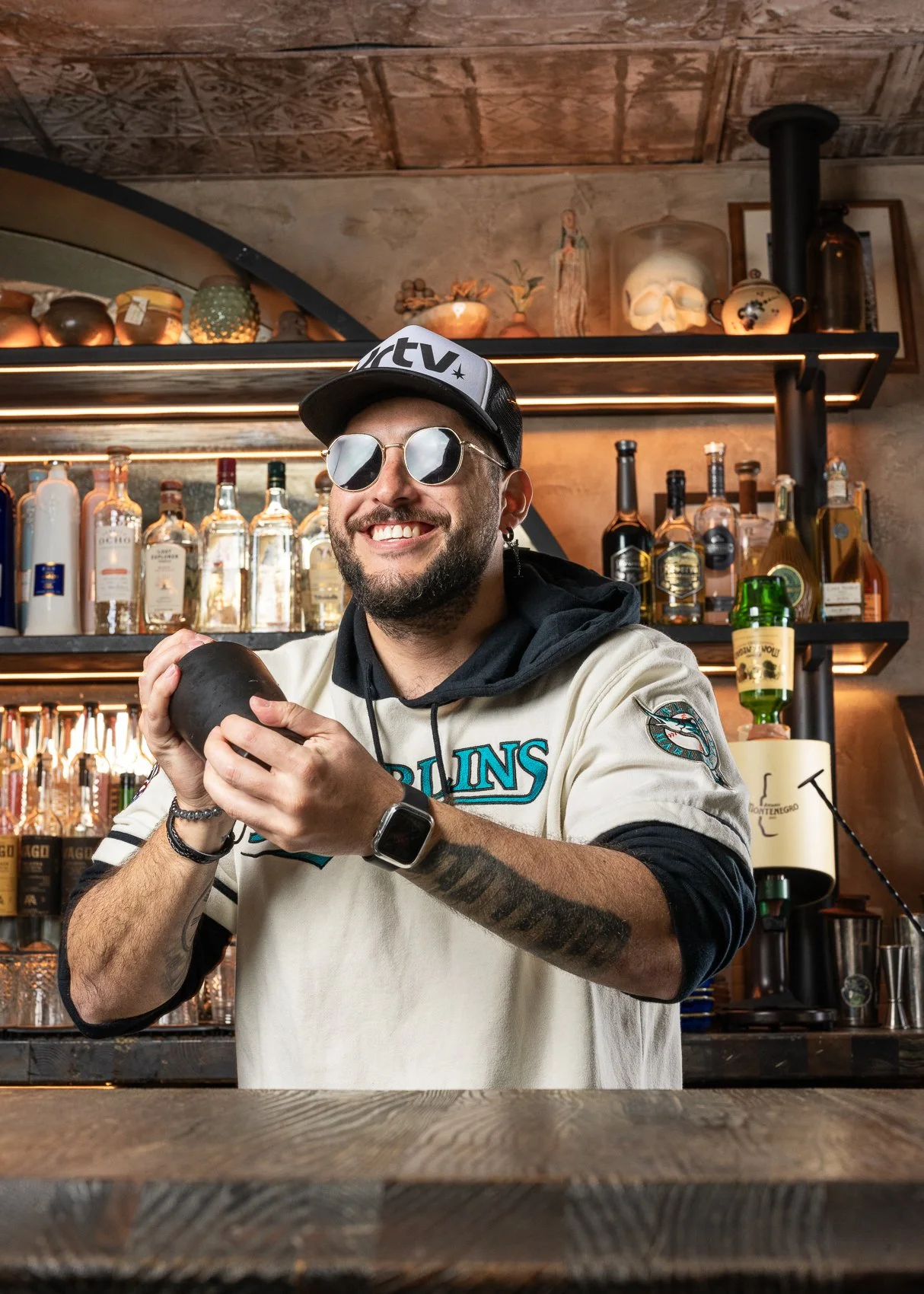 Bartender Stefano smiling while shaking a cocktail at the mezcaleria bar inside Bodega Taqueria y Tequila in Coconut Grove, wearing a Miami Marlins jersey and sunglasses.