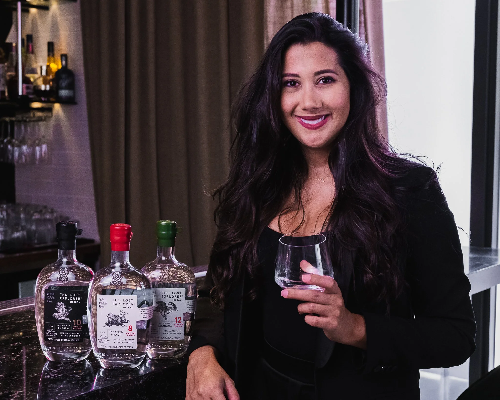 Portrait of Helena José, brand representative for The Lost Explorer Mezcal, holding a tasting glass beside a display of mezcal bottles.