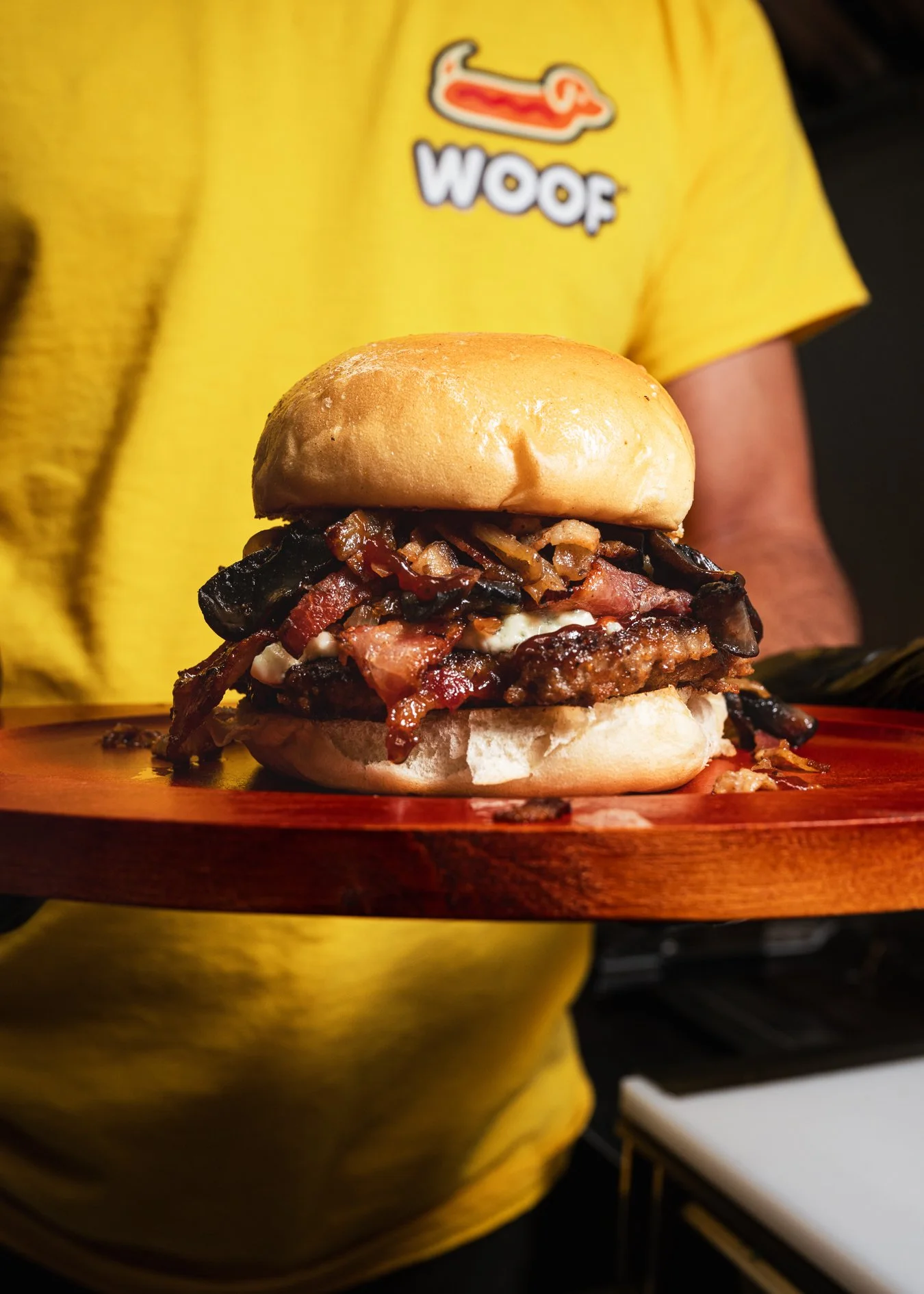 Mushroom bacon cheeseburger stacked with grilled onions and melted cheese, photographed for Woof Hotdog food truck in Doral with a staff member wearing the Woof logo shirt in the background.